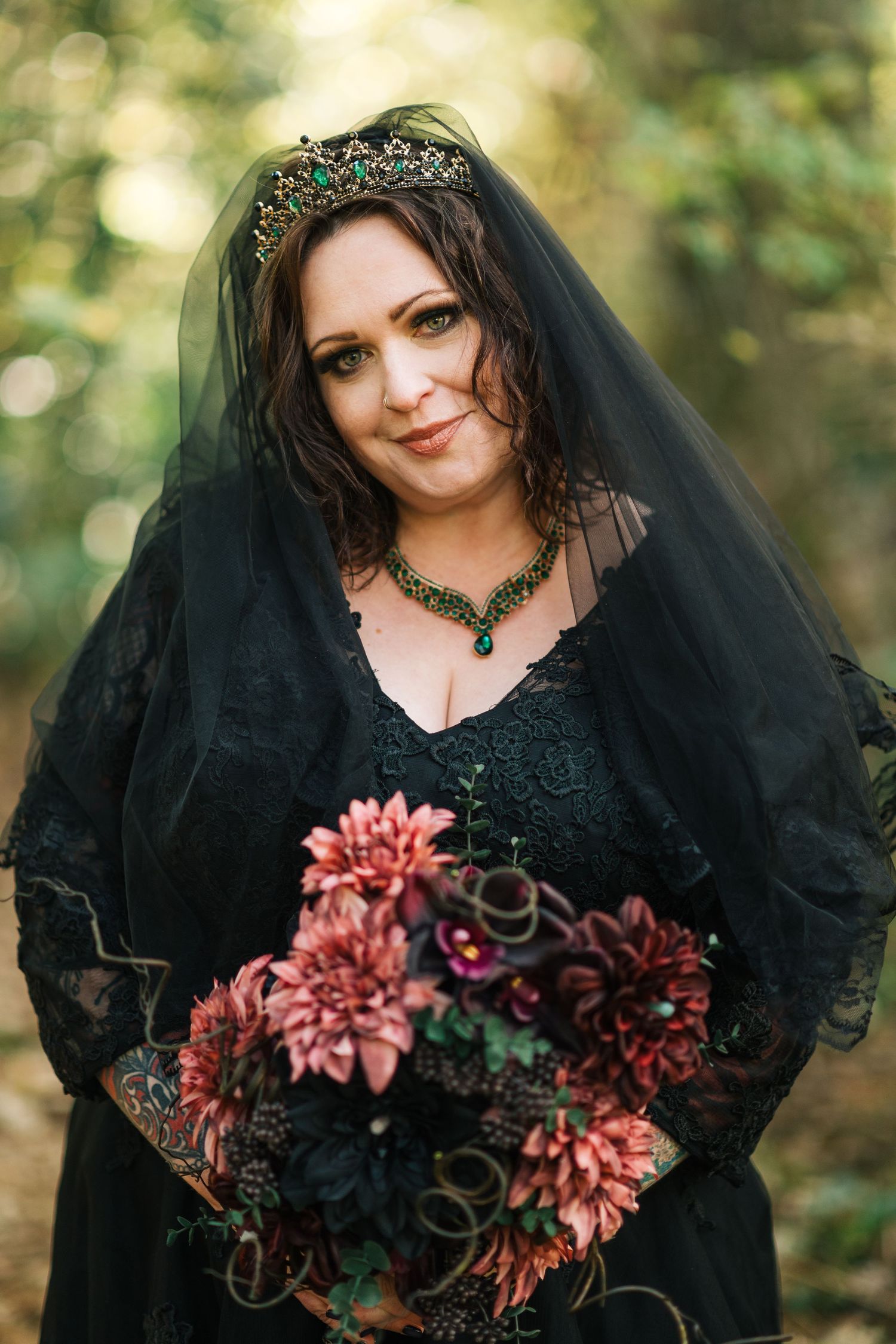 Gothic bride in black lace gown and crown veil holds dark floral bouquet of burgundy dahlias in forest setting.
