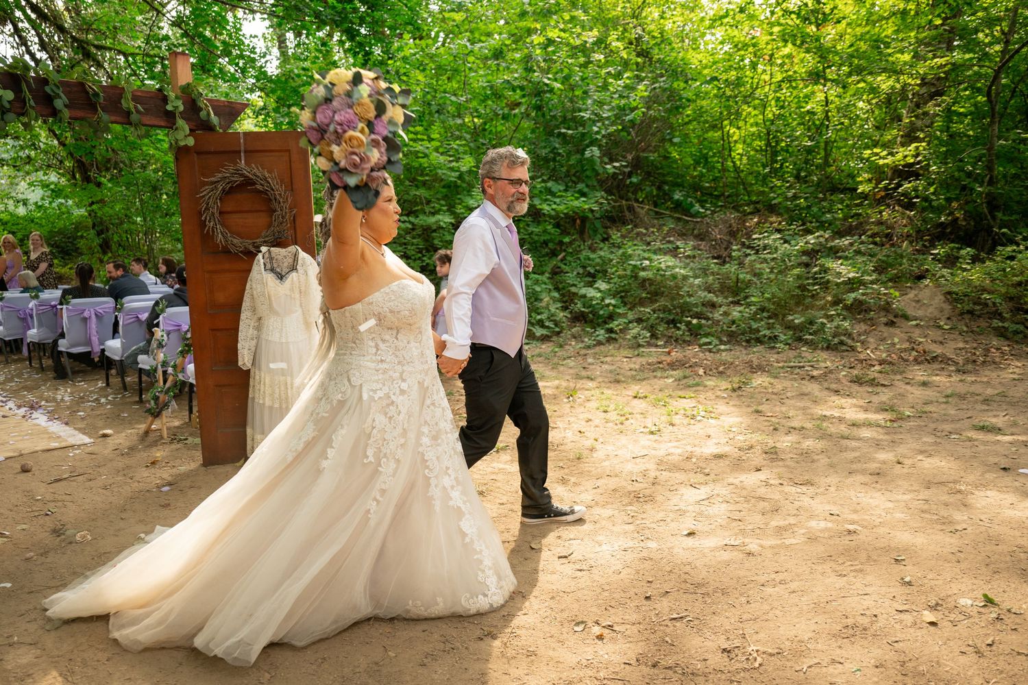 Bride in lace gown and groom in pink shirt walk hand in hand past rustic wooden door at outdoor wedding.