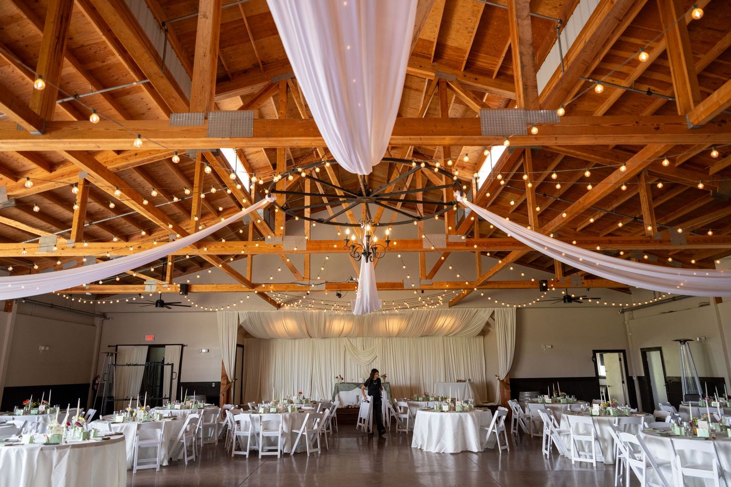 Elegant wedding reception hall with wooden beam ceiling, string lights, white draping, and round tables with white chairs.