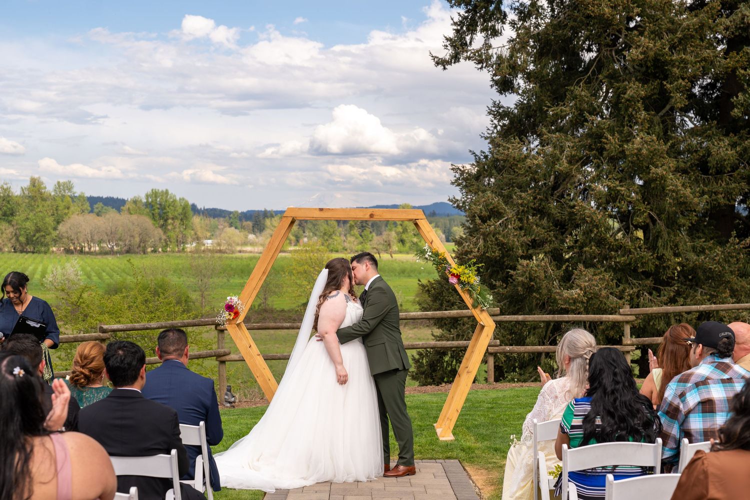 Bride and groom share first kiss under hexagonal wooden arch at outdoor wedding ceremony with scenic green valley backdrop.