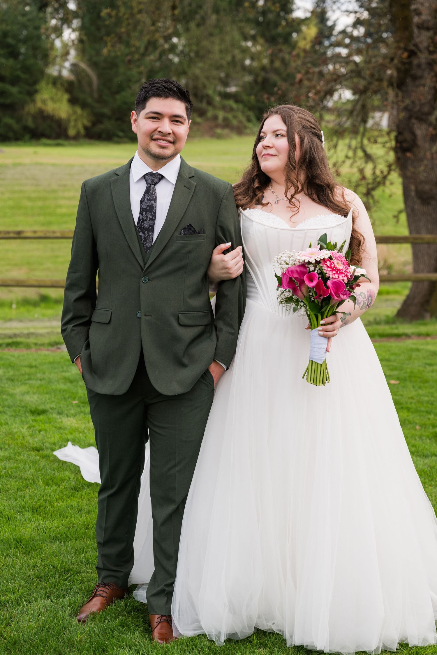 A bride and groom pose outdoors on green grass, she holds pink bouquet in white gown, he wears olive suit.