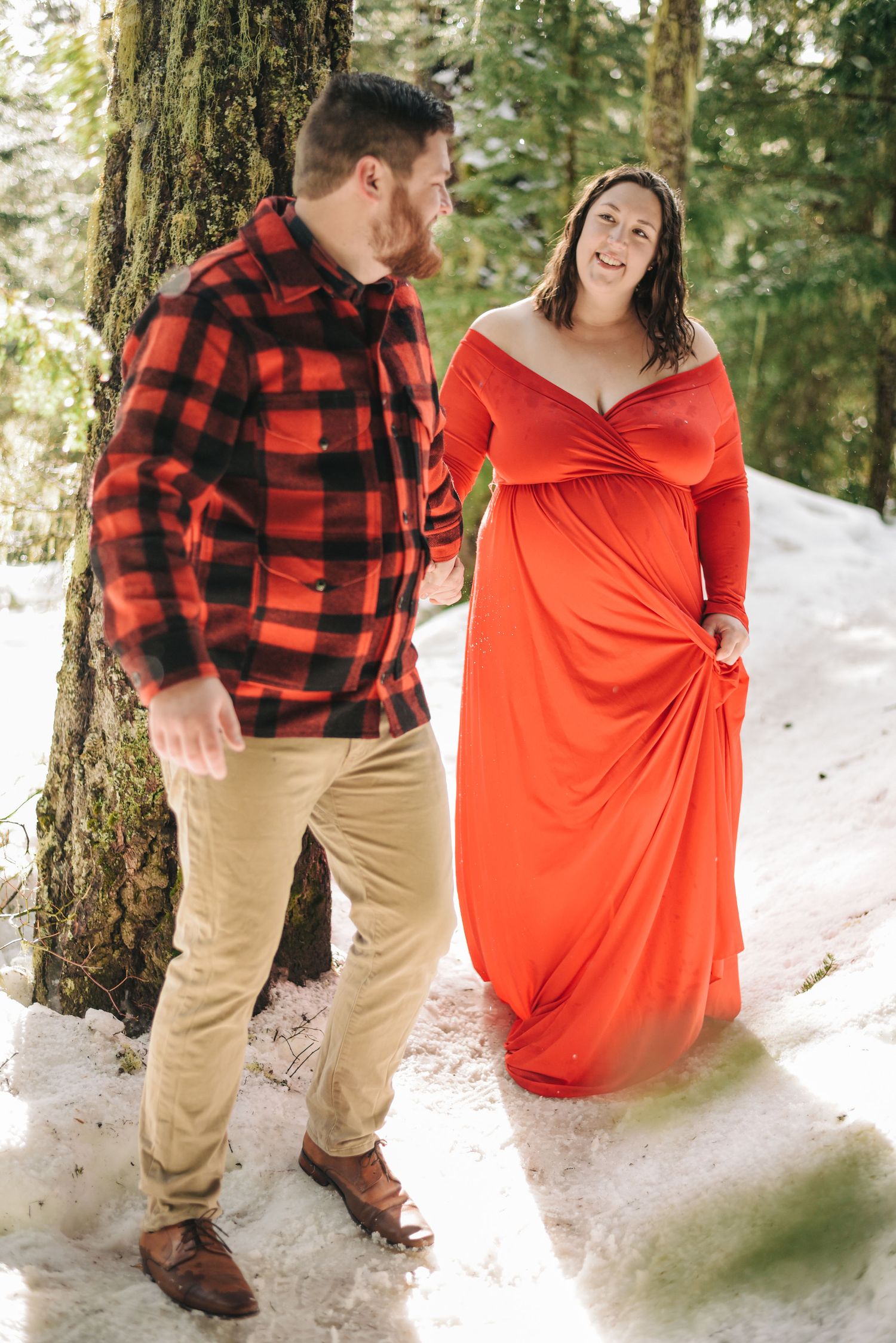 Playful winter couple photos with red dress and buffalo plaid in snowy forest.