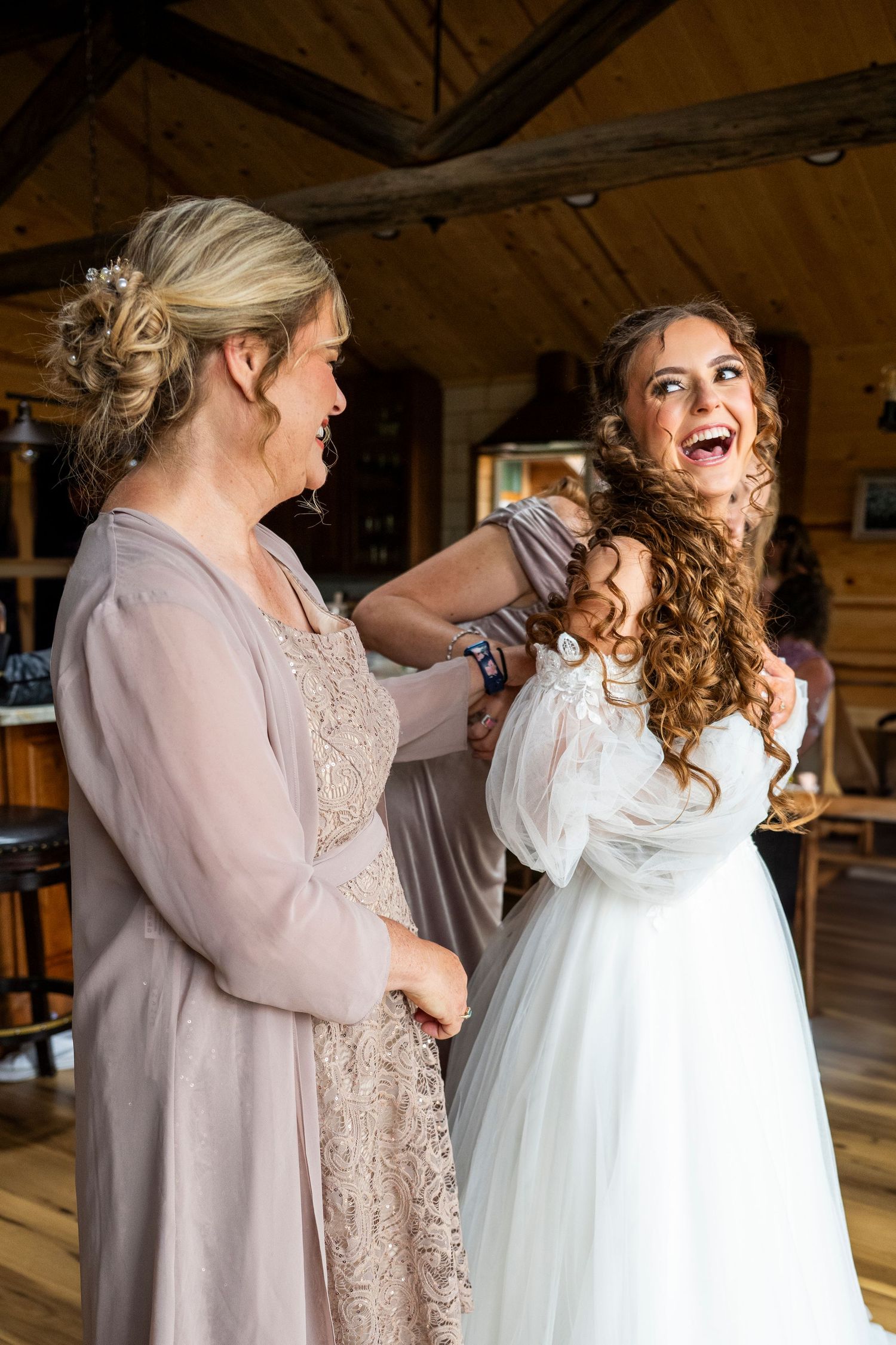 Bride laughing joyfully as bridesmaid helps her into her white wedding gown inside a rustic wooden venue.