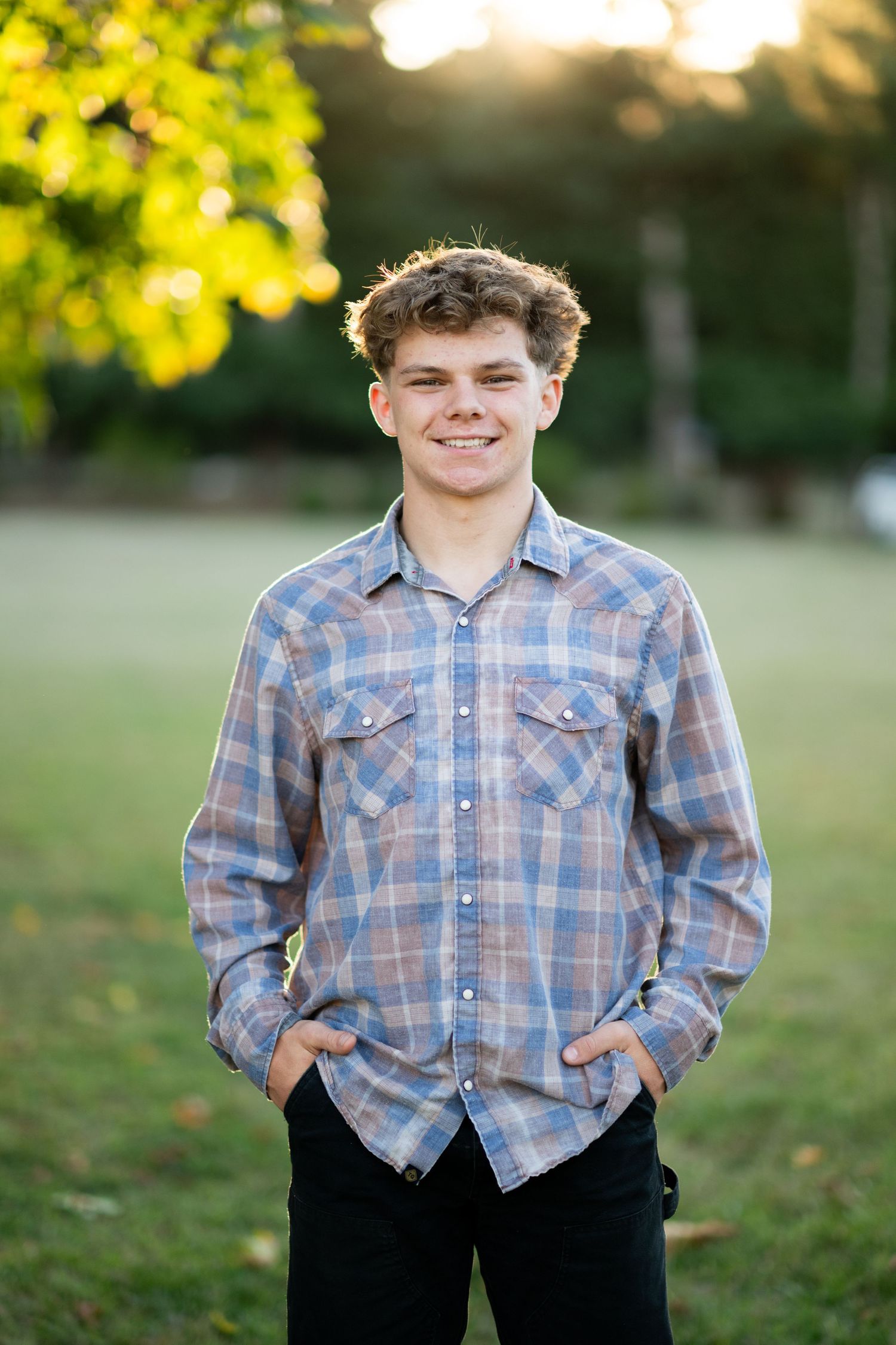 Smiling teenage boy in a plaid shirt standing with hands in pockets in a sunlit outdoor field with trees.