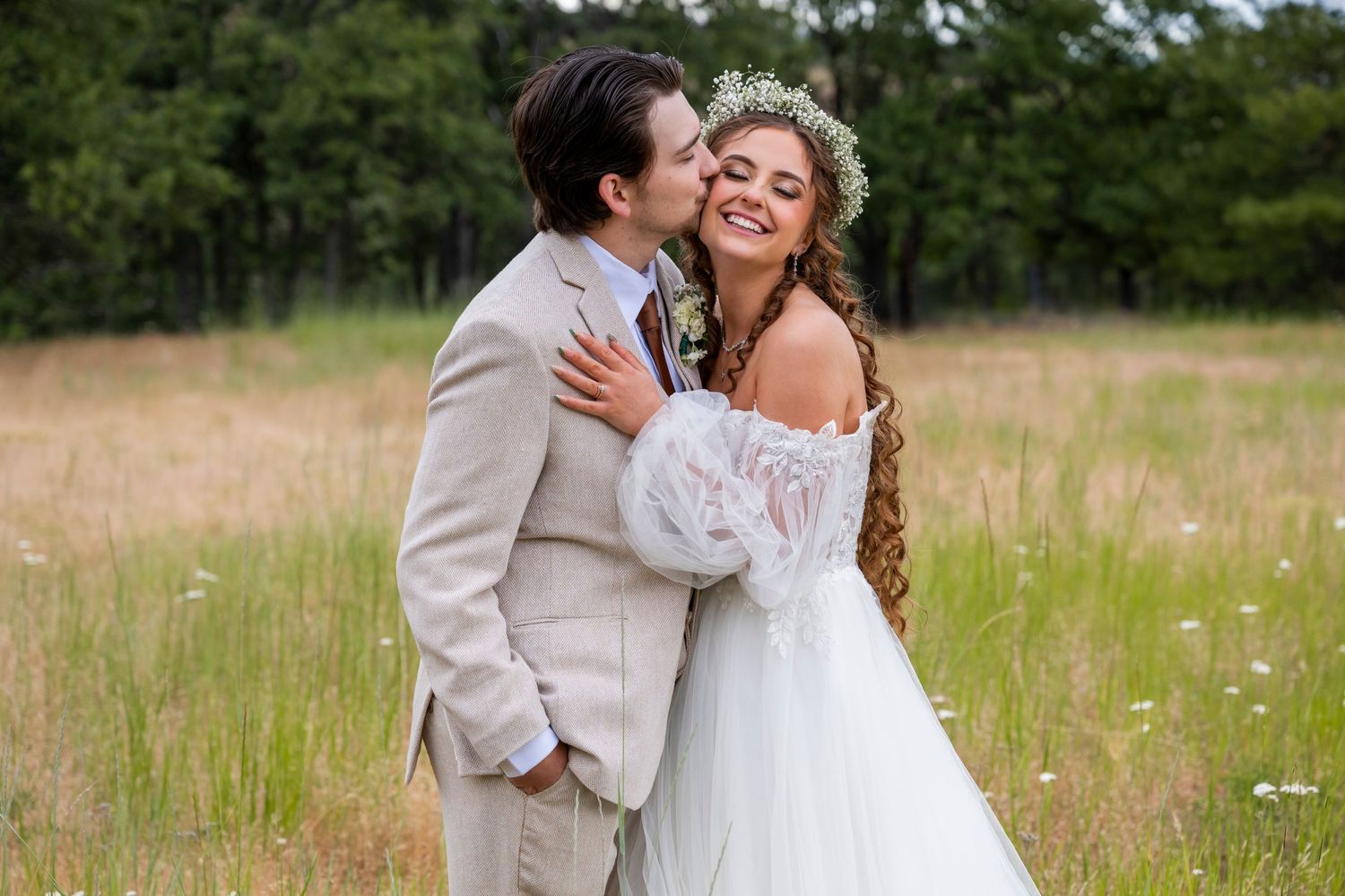 Groom in tan suit kissing smiling bride wearing baby's breath crown in a sunlit meadow with wildflowers.