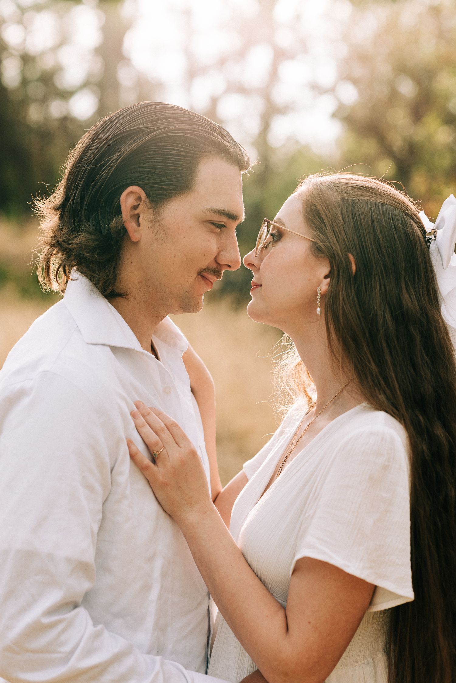 A couple in white attire share an intimate nose-to-nose moment in a golden field.
