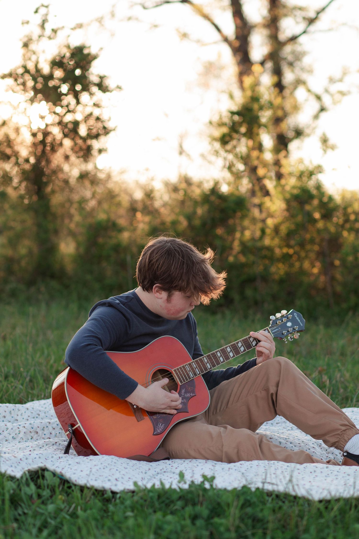 Young man playing acoustic guitar on a blanket in a green field at sunset.