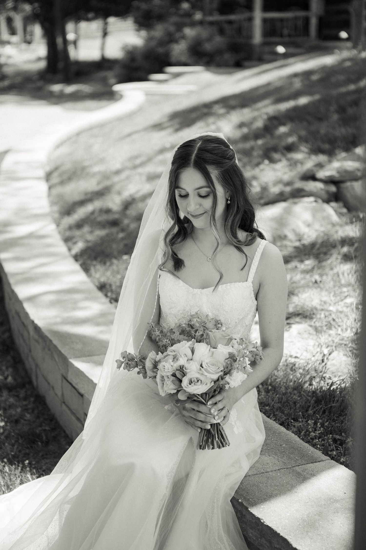 Bride in white gown holds bouquet, looking down gracefully outdoors in black and white wedding portrait.