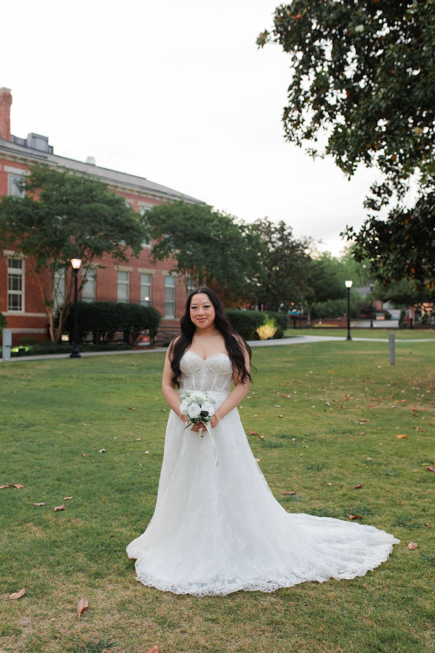 Bride in a white lace gown holding a bouquet stands on green lawn before a brick building.