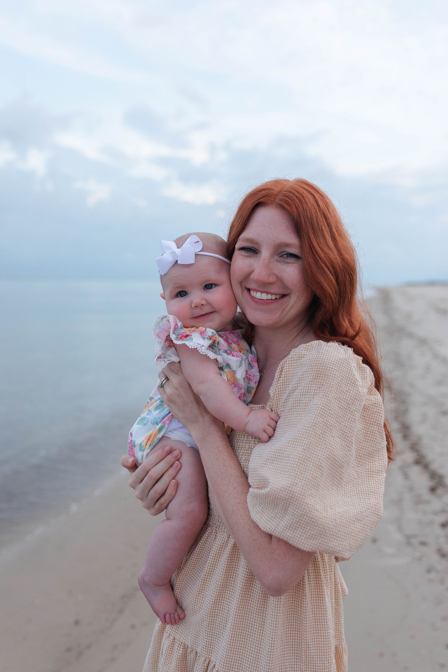 Red-haired woman in beige dress holds smiling baby girl with white bow headband on sandy beach.