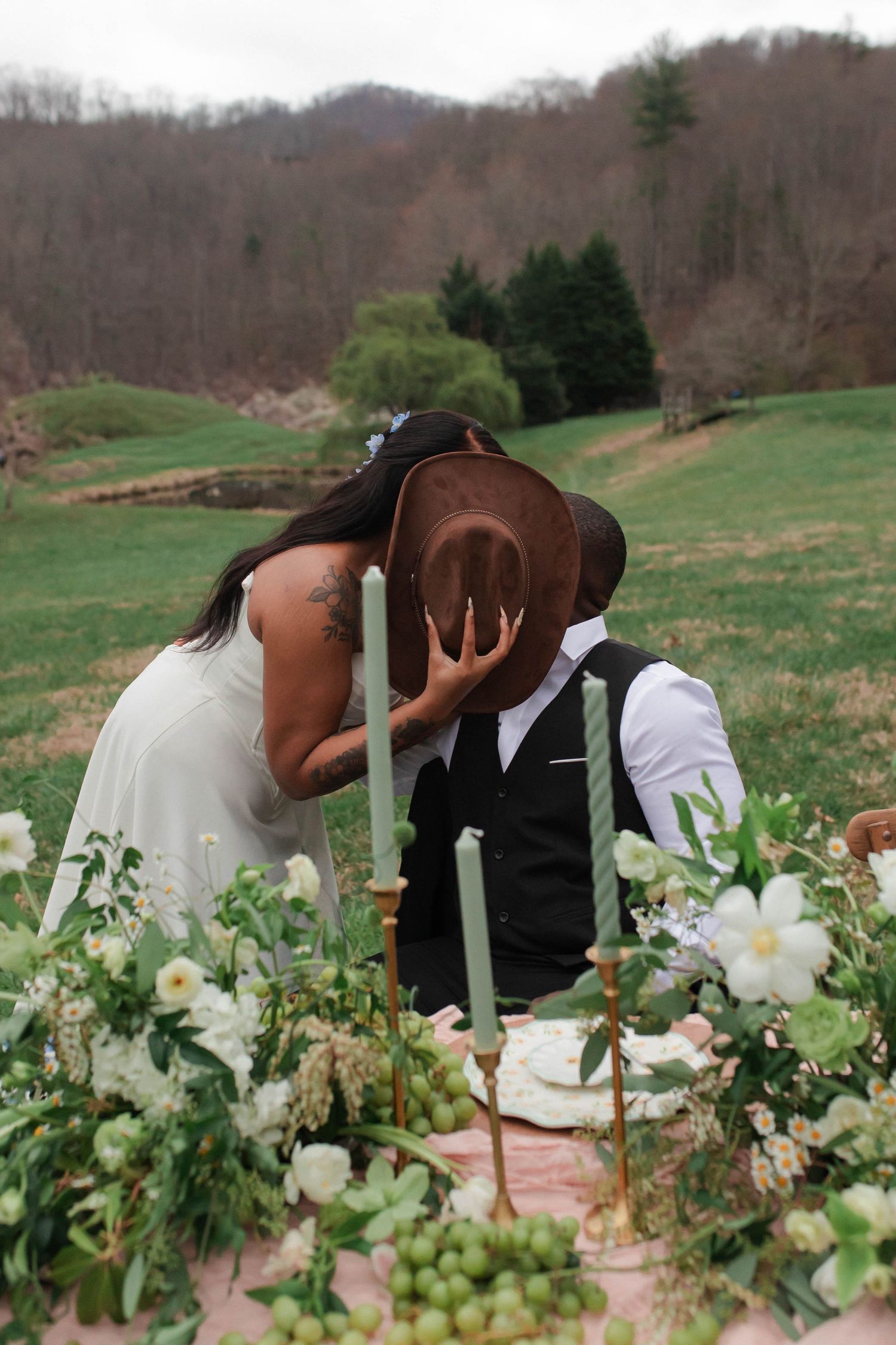 Bride and groom share a kiss hidden behind a brown cowboy hat at an outdoor floral wedding tablescape after eloping in Nashville.