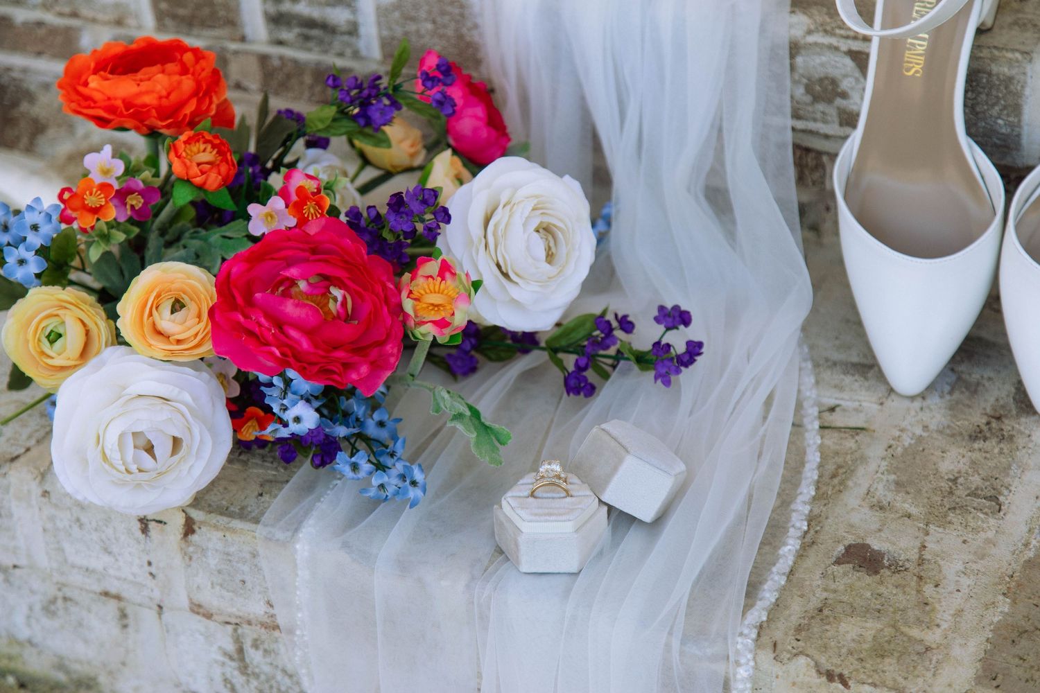 Colorful wedding bouquet with ranunculus and wildflowers beside white heels and ring box on stone surface.