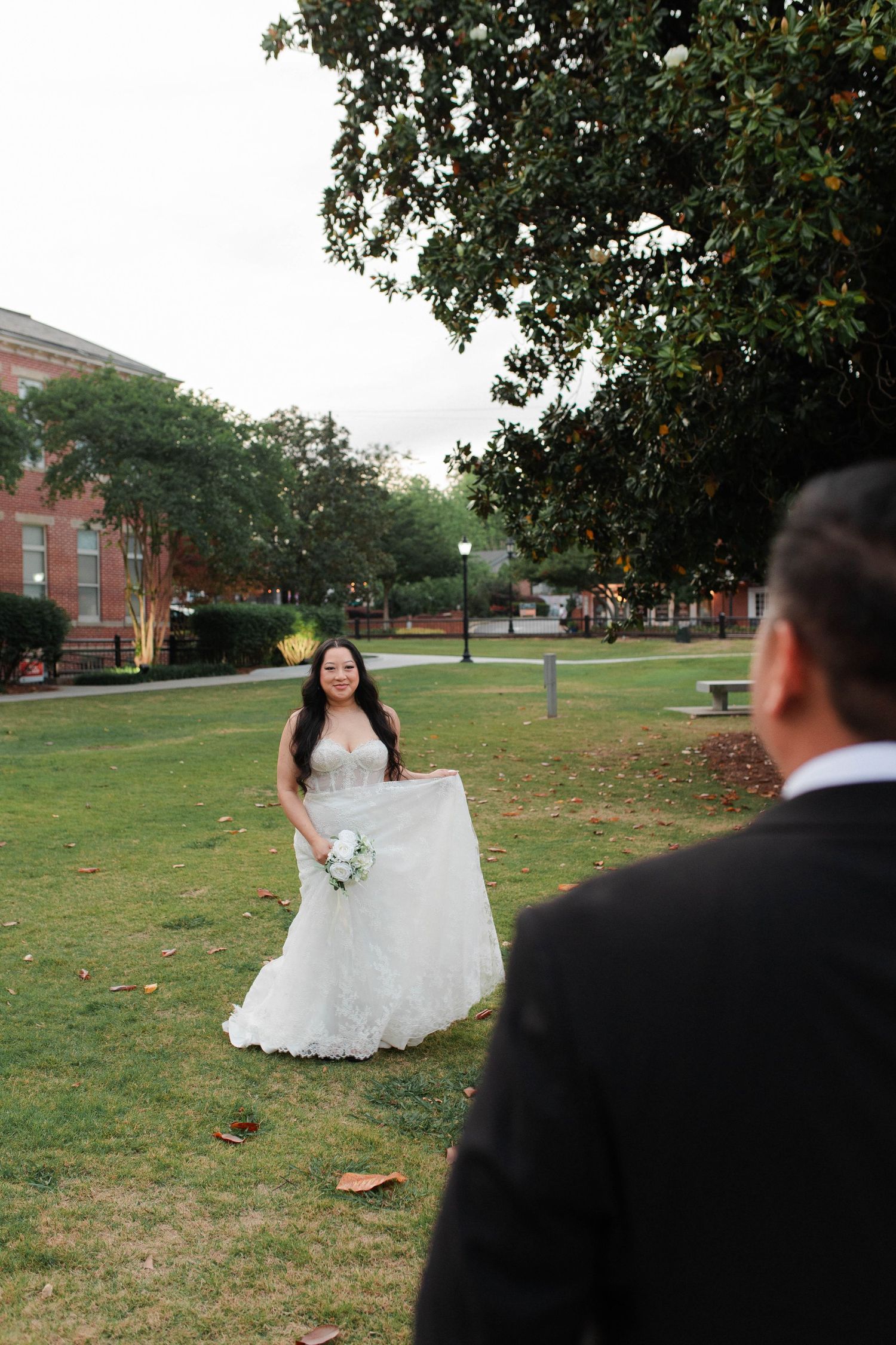 Bride in white lace gown smiling and holding her dress on a green lawn during wedding photos.