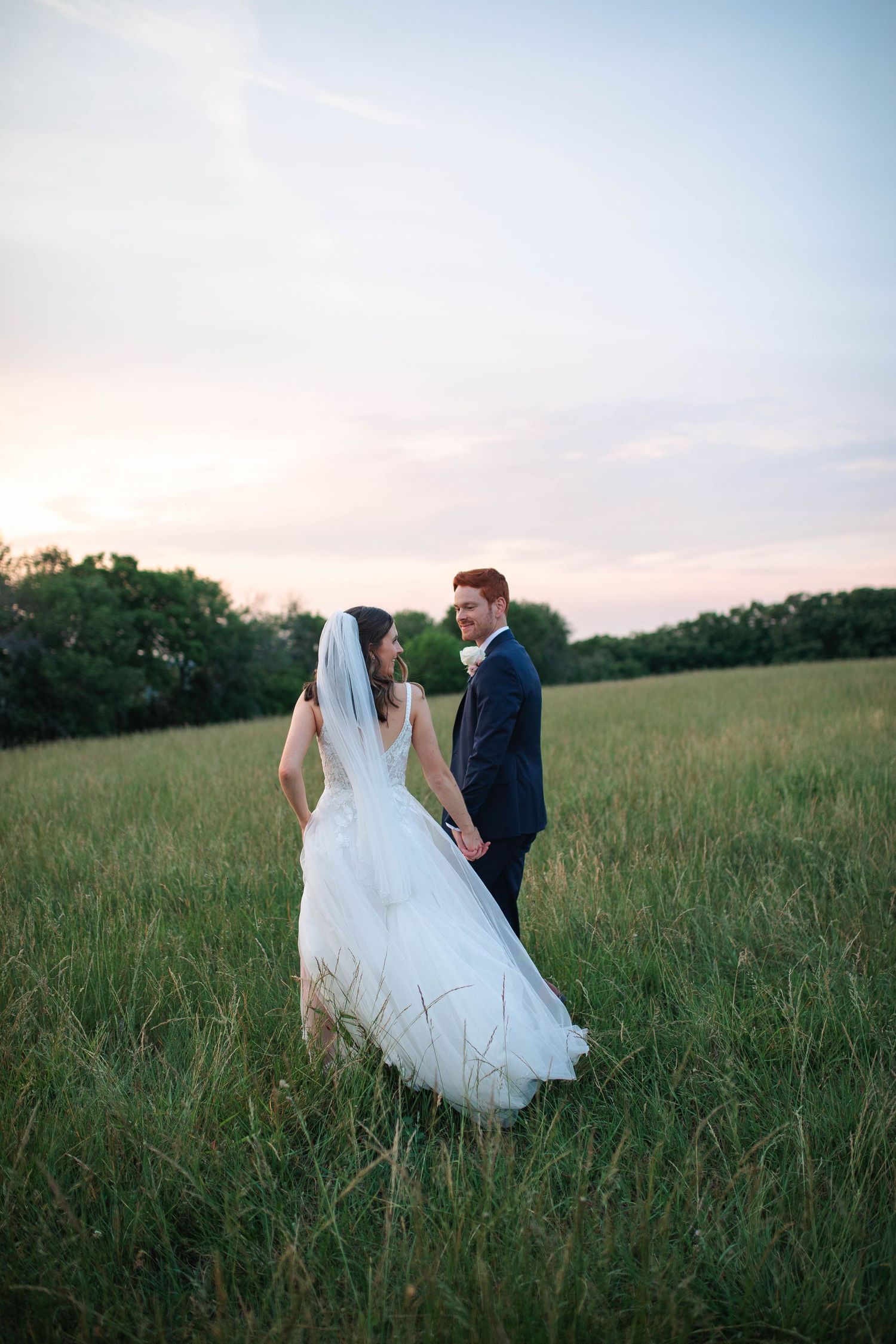 Bride and groom holding hands walking through green meadow at sunset, bride in white gown with veil, groom in navy suit.