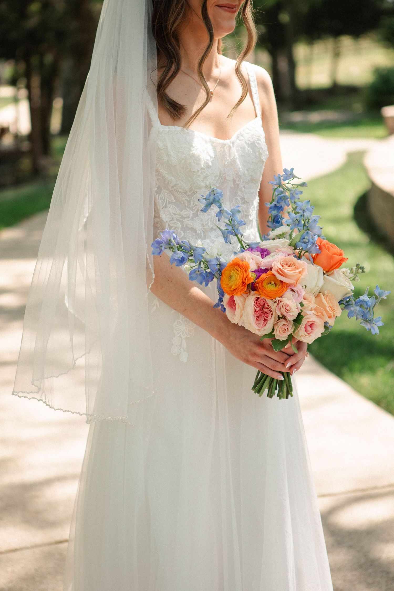 Bride in white lace gown and veil holding colorful bouquet with orange, pink, and blue flowers outdoors.