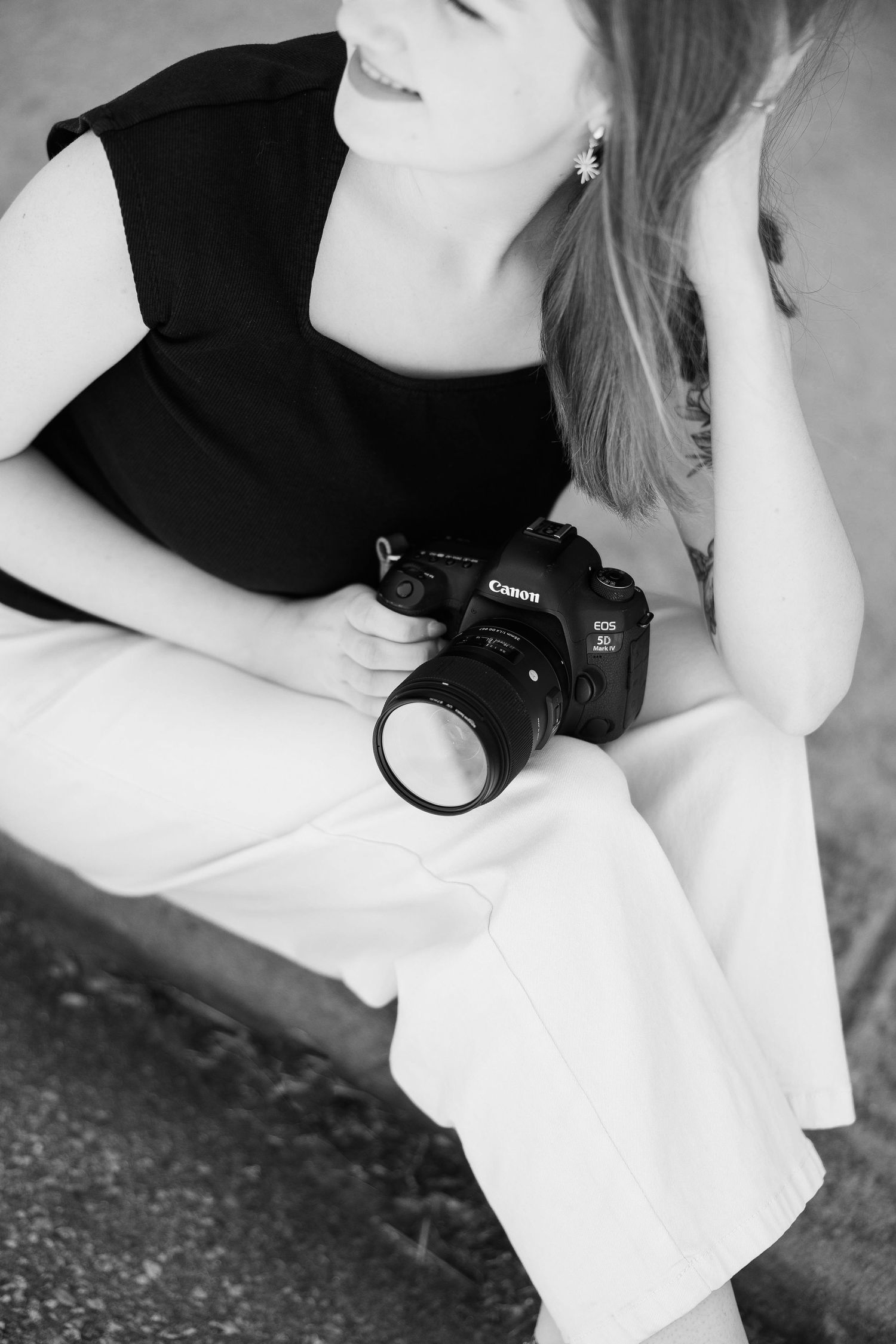 Black and white photo of a smiling woman sitting outdoors holding a Canon EOS DSLR camera, wearing a black top and white pants.