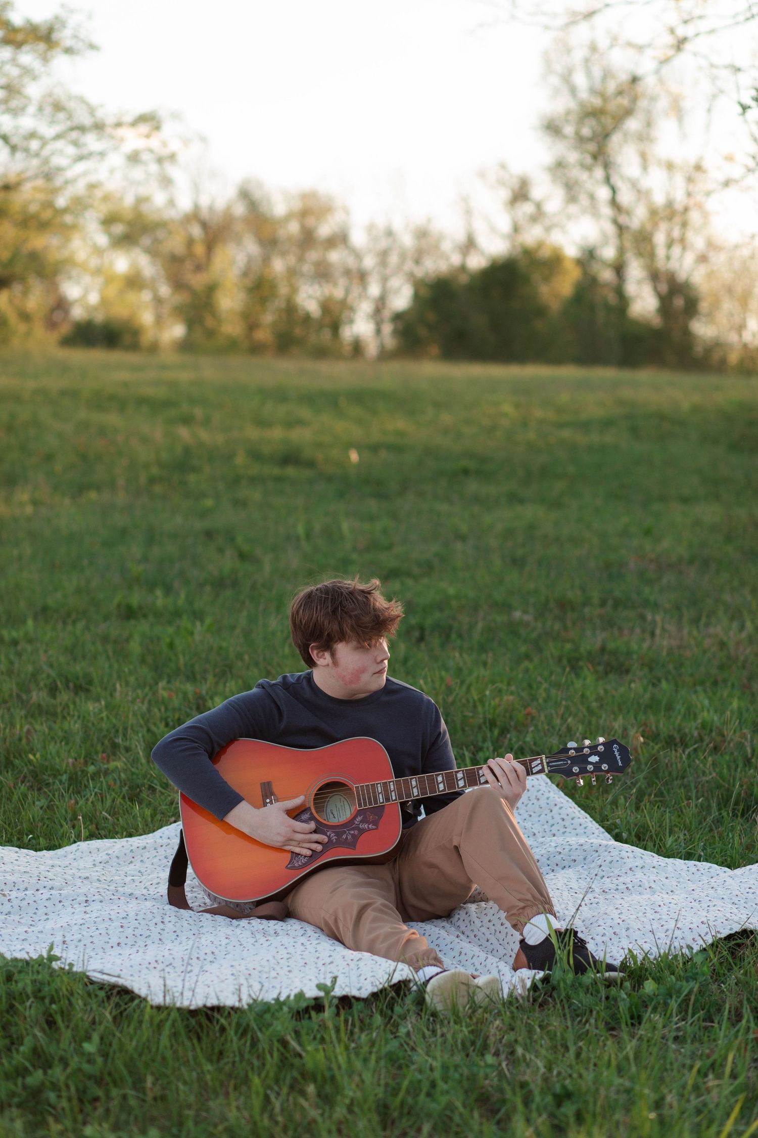 Young man playing acoustic guitar on a blanket in a green field during golden hour sunset.
