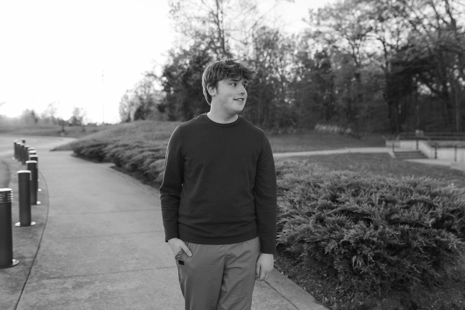 Young man in dark sweater smiling while walking along a park pathway surrounded by lush greenery.