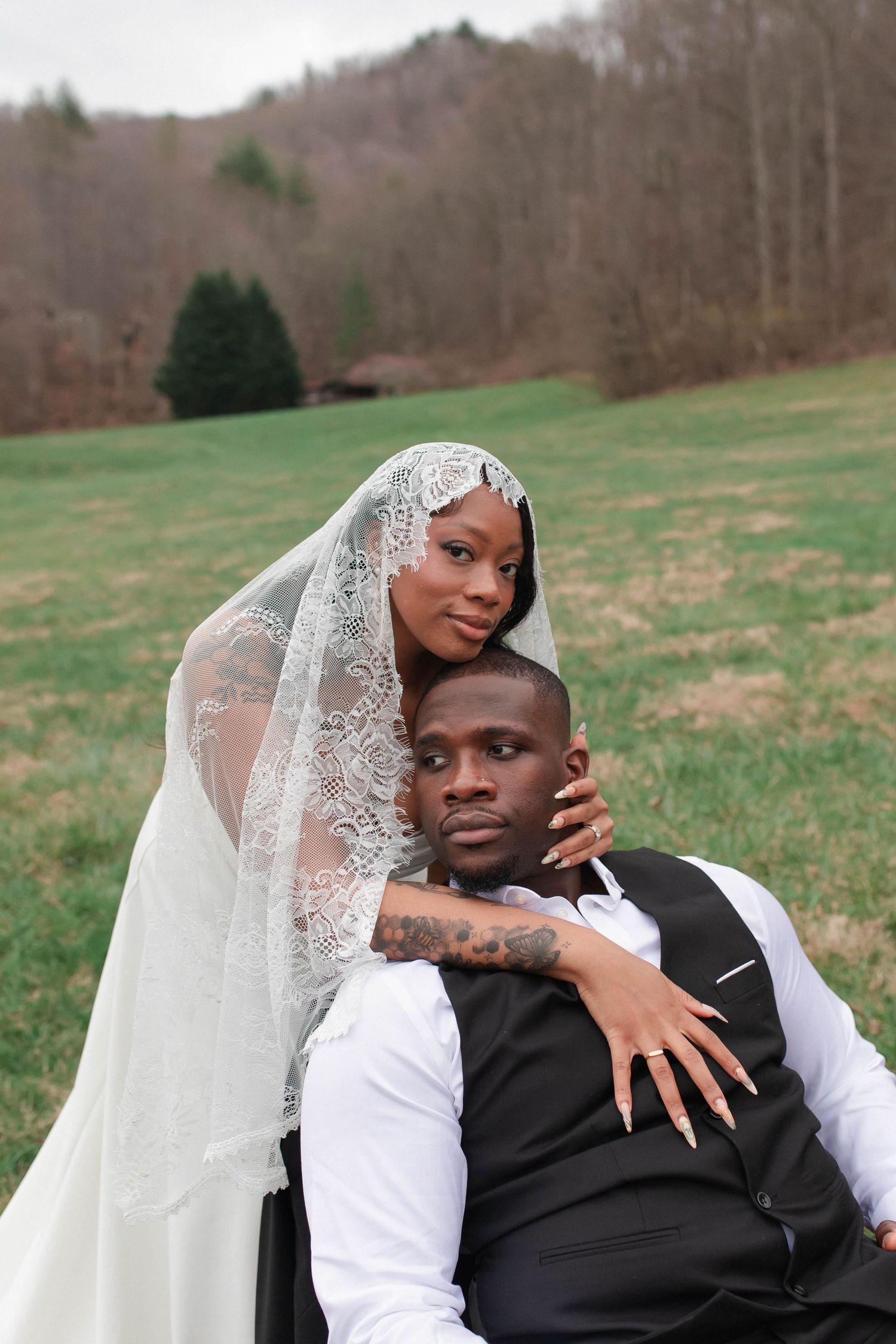 A bride in lace veil embraces groom in black vest outdoors on green grass at a wedding portrait session.