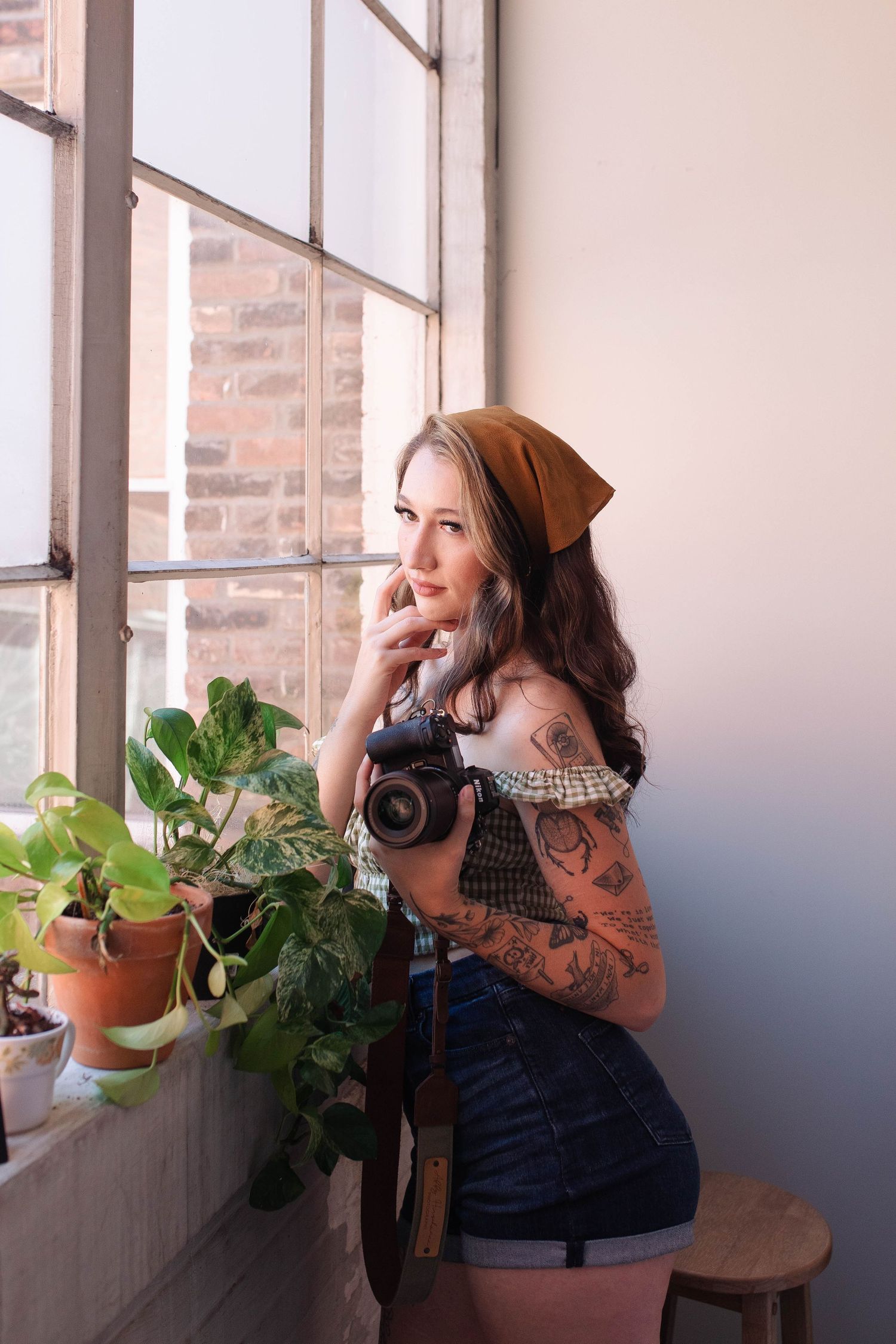 Young tattooed woman holding a camera poses by a sunlit window with potted plants, wearing denim shorts and a headscarf.