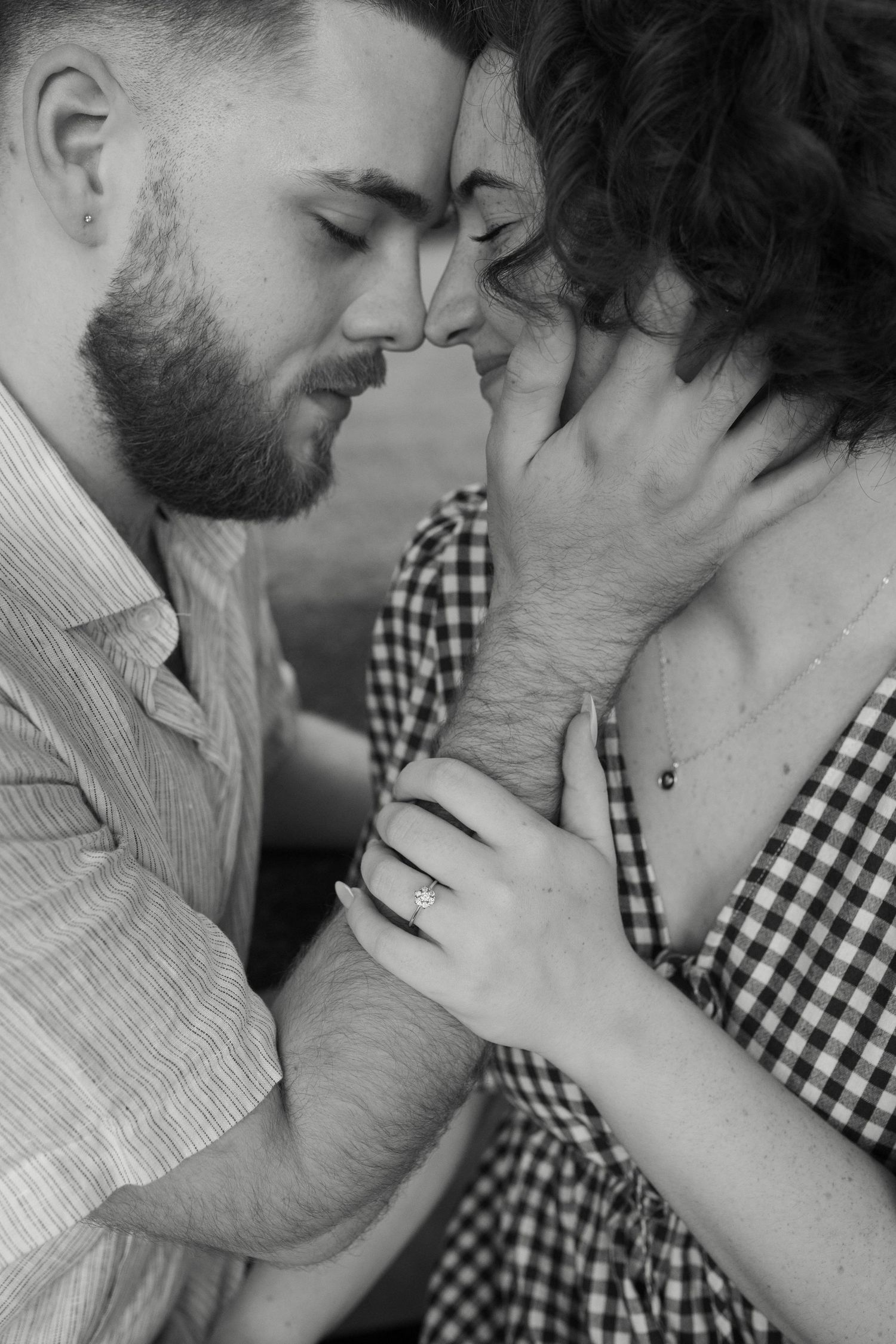 Black and white photo of a romantic couple with foreheads touching, man tenderly holding woman's face, wearing engagement ring.