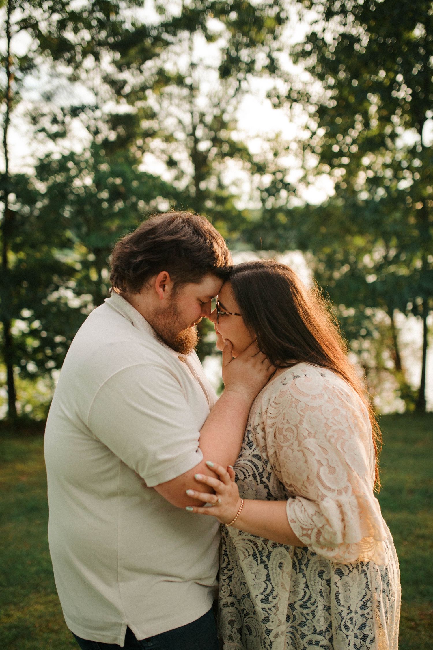 Couple sharing a romantic forehead touch outdoors, surrounded by lush green trees in soft golden light.
