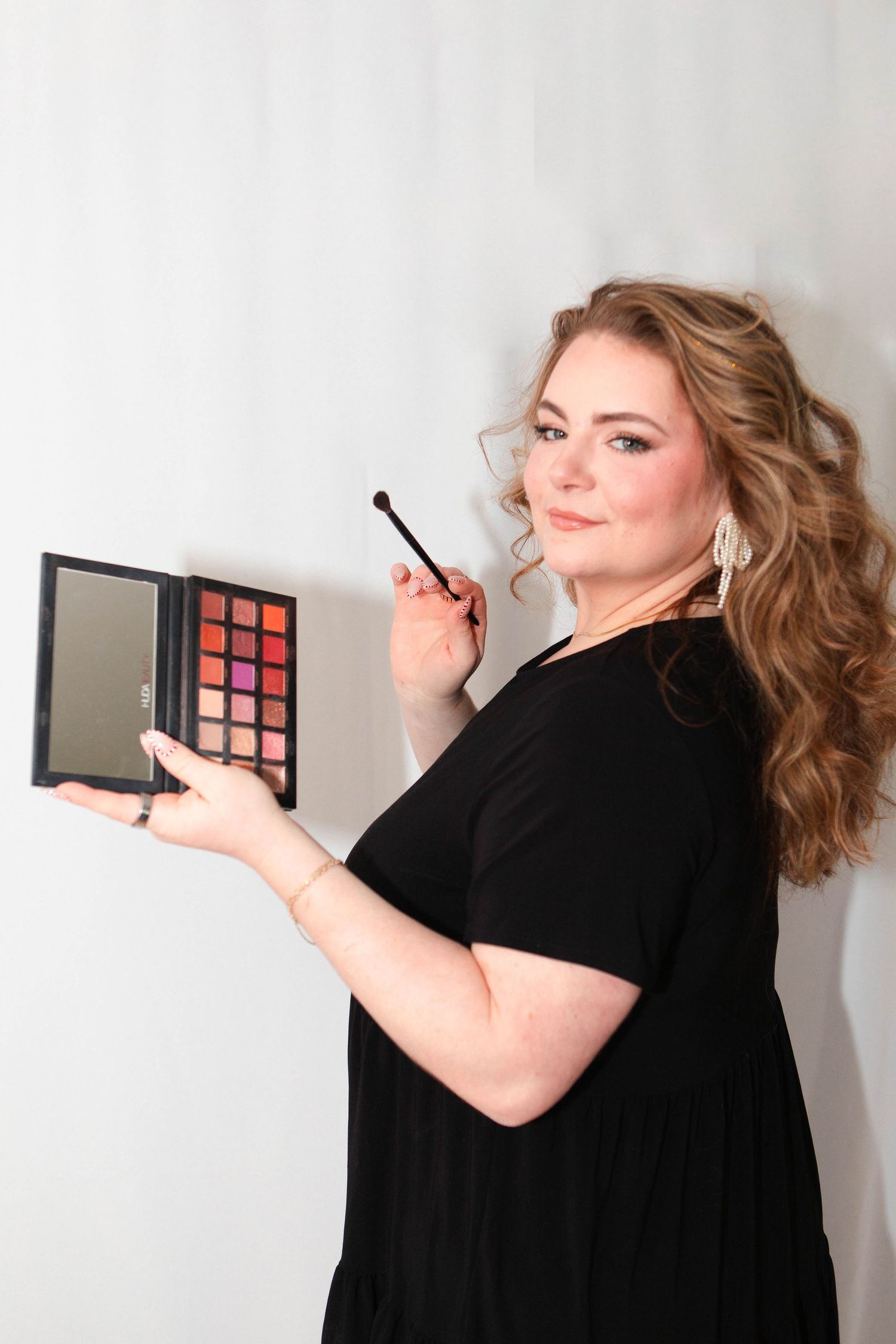 A plus-size woman in a black dress holds a colorful eyeshadow palette and brush, posing as a makeup artist.