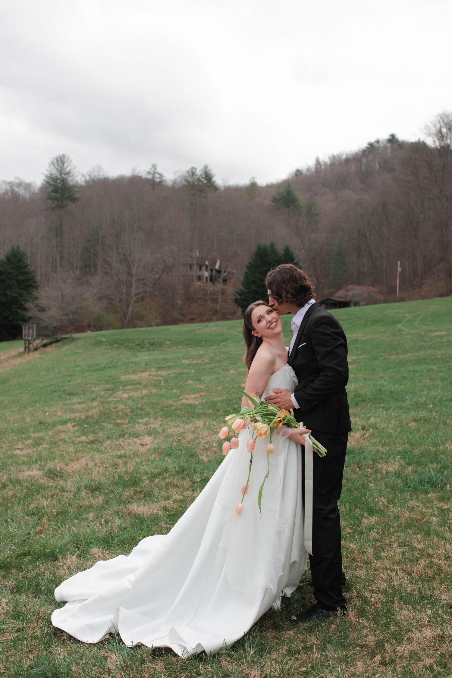 Groom in black suit kisses bride's head as she smiles holding tulip bouquet on a green hillside after eloping in Nashville.