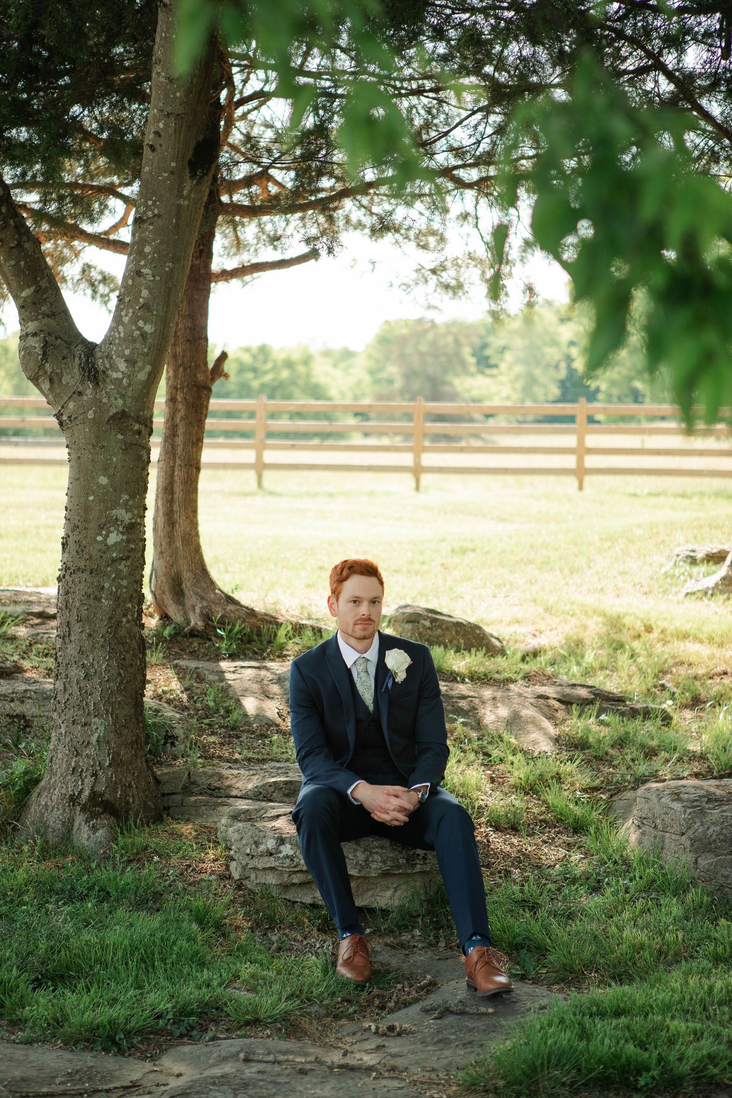 A groom in a navy suit with white boutonniere sits on stone steps beneath a tree at an outdoor farm wedding venue.