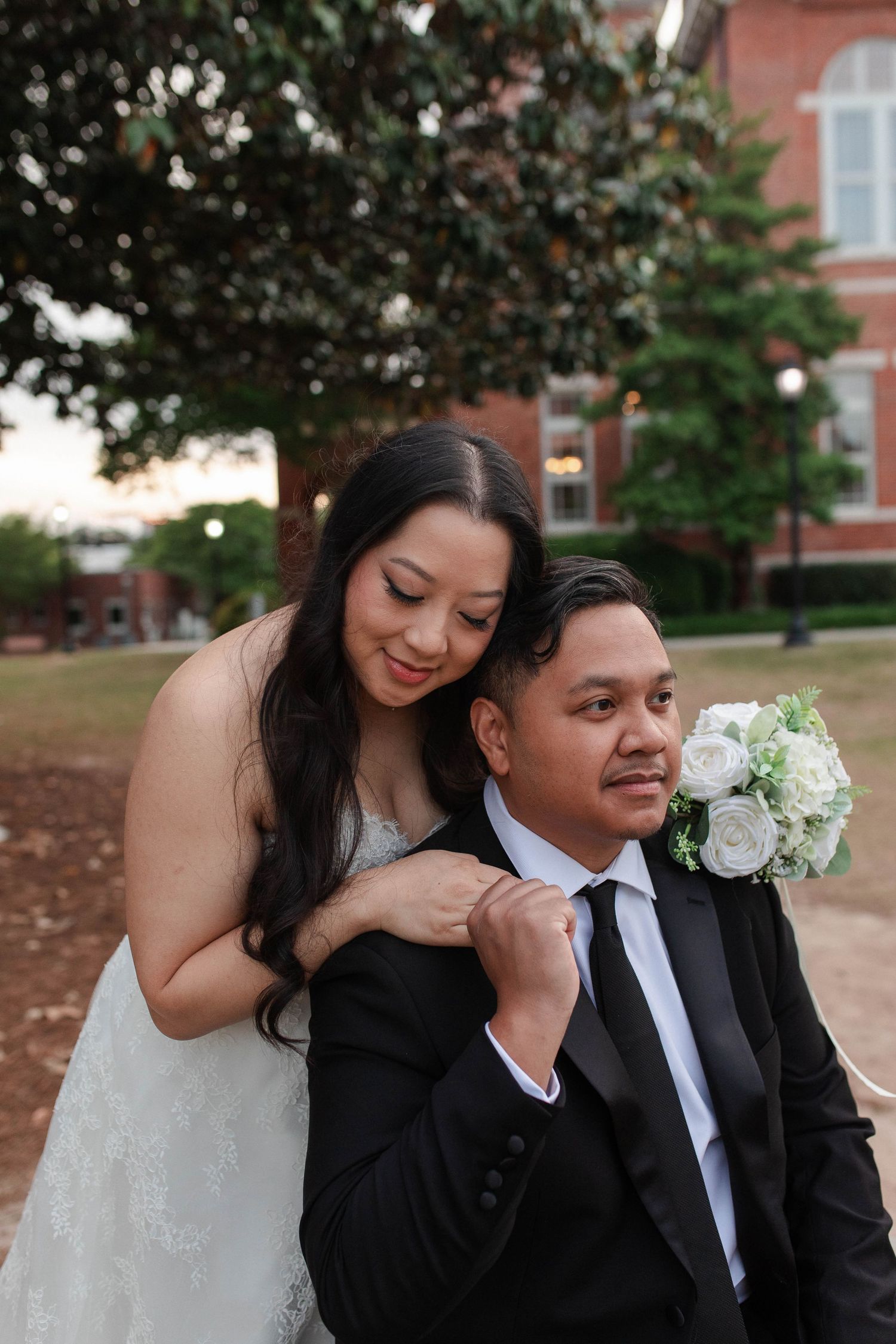A bride in a white gown embraces her groom in a black tuxedo outdoors, holding a white rose bouquet near a brick building.