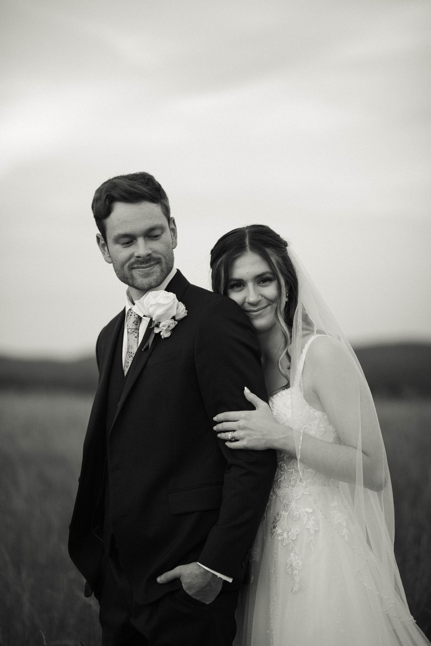 Black and white wedding portrait of bride and groom posing in a field, bride leaning on groom's shoulder.