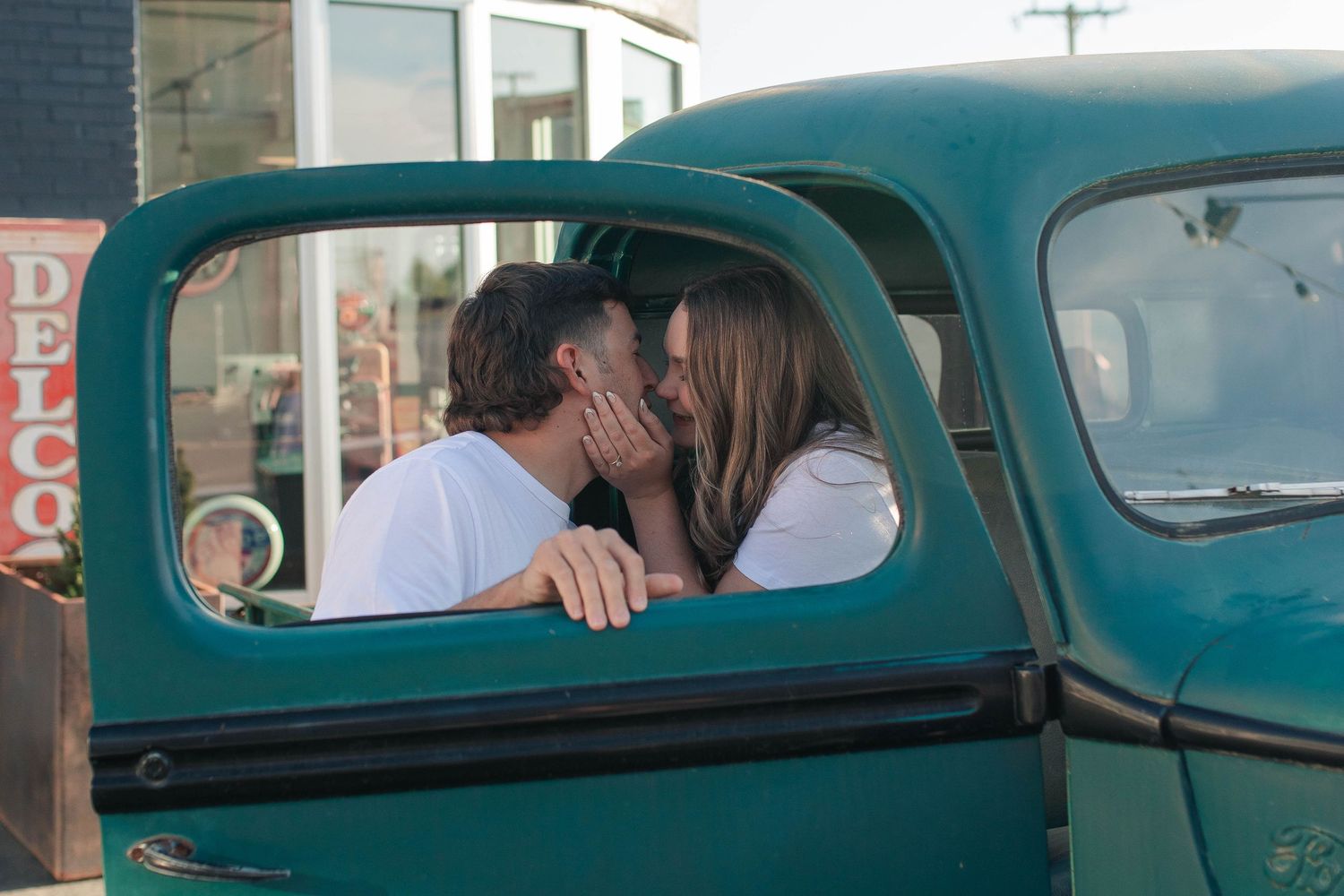 Couple sharing a romantic kiss inside a vintage teal green truck parked outside a retro shop.