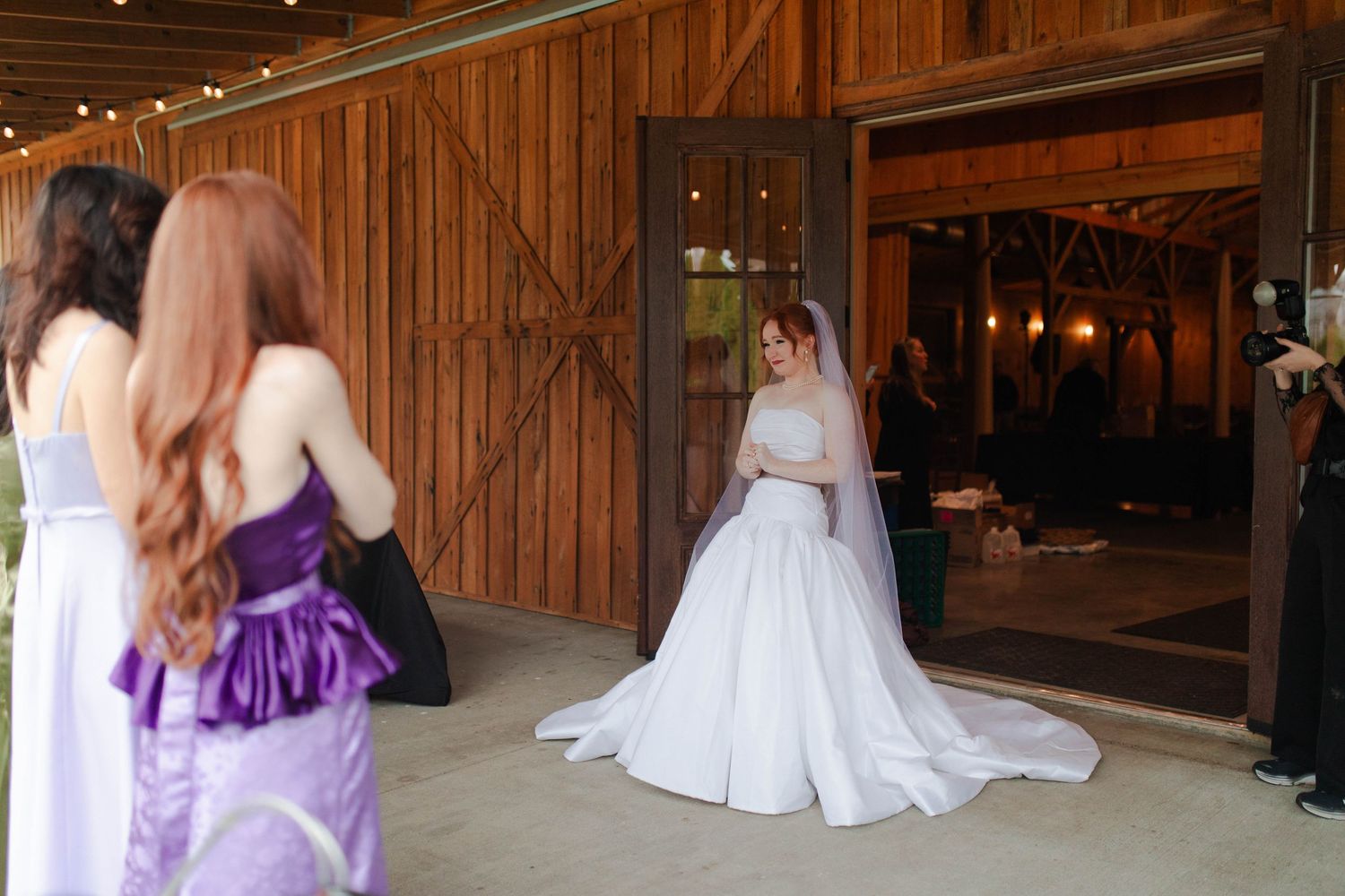 Nashville micro wedding bride in white gown with veil poses near barn doors while bridesmaids in purple dresses look on.