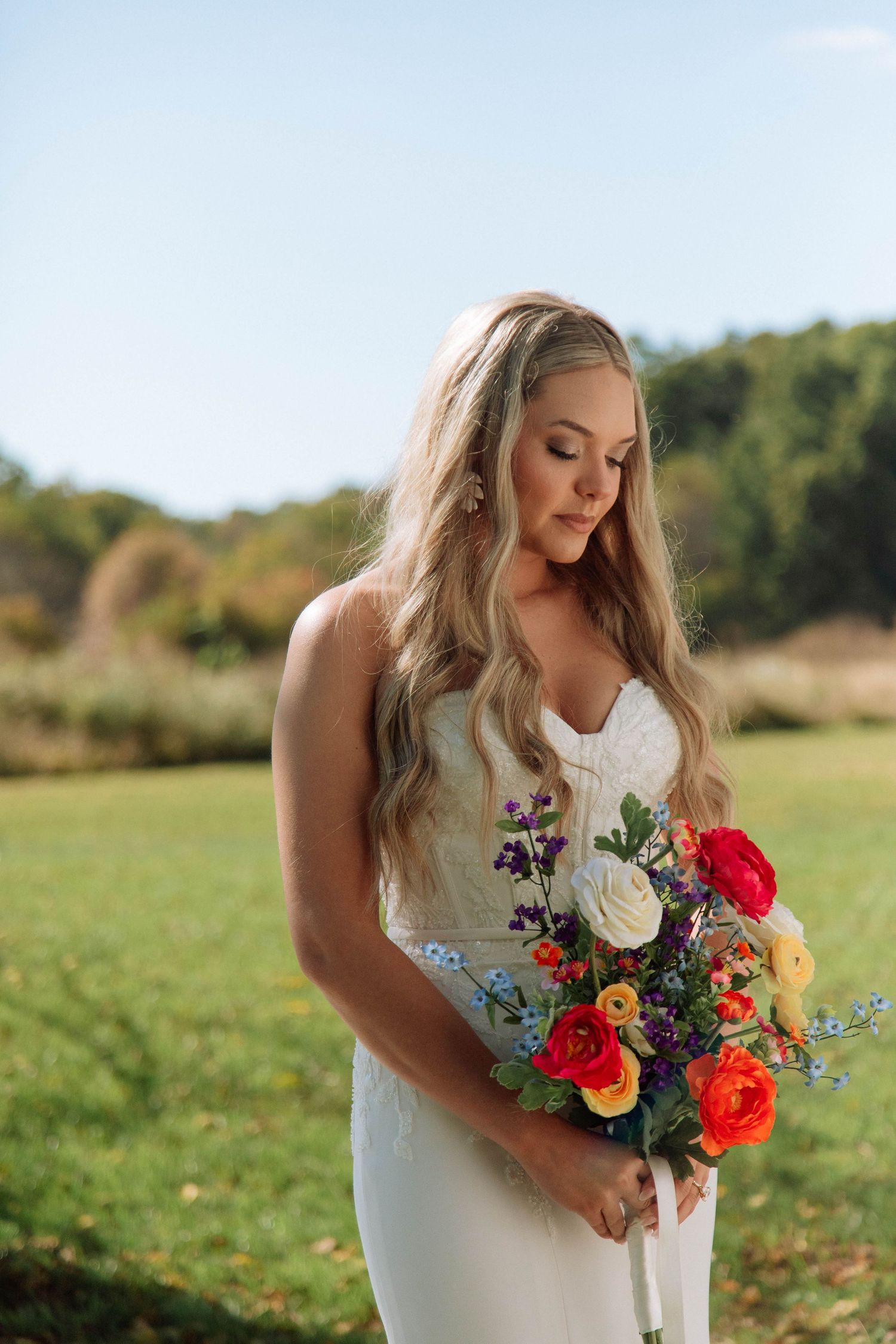 Blonde bride in white fitted gown gazing down at vibrant orange, red, and white bouquet in sunny field after eloping in Nashville.