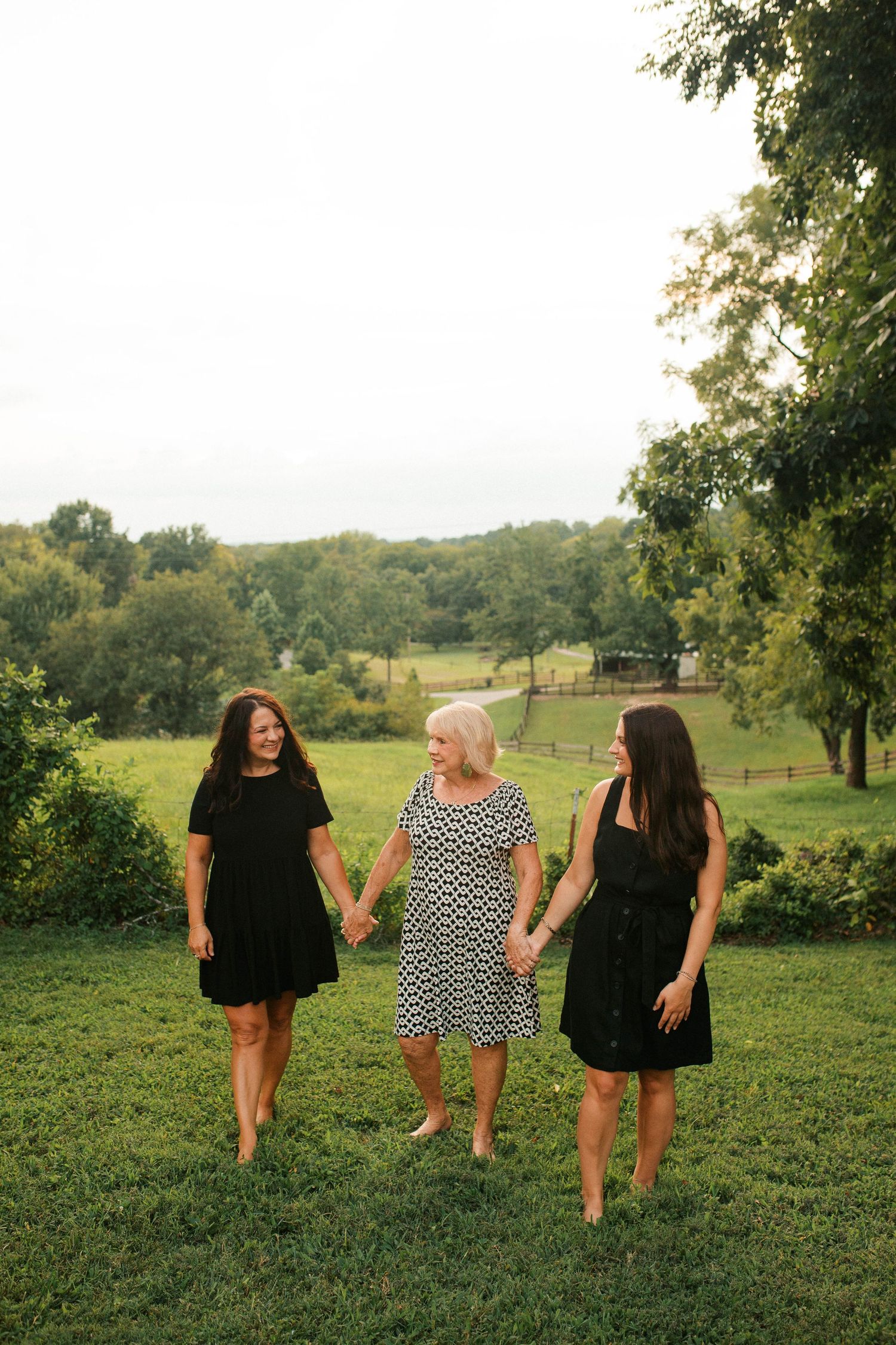 Three women in black and white dresses hold hands while walking on green grass with trees in background.