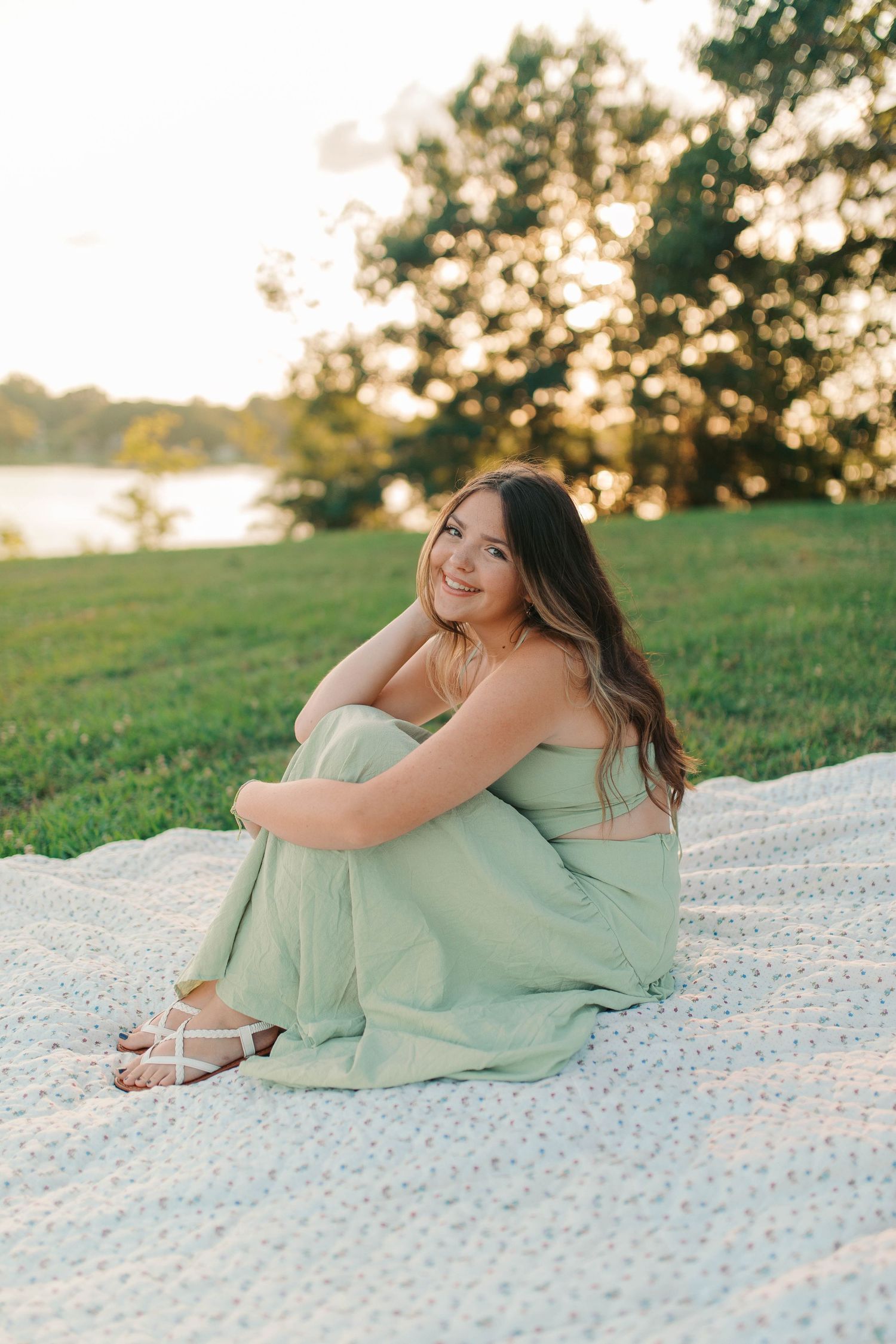 Young woman in mint green jumpsuit sitting on blanket outdoors at golden hour near lake.