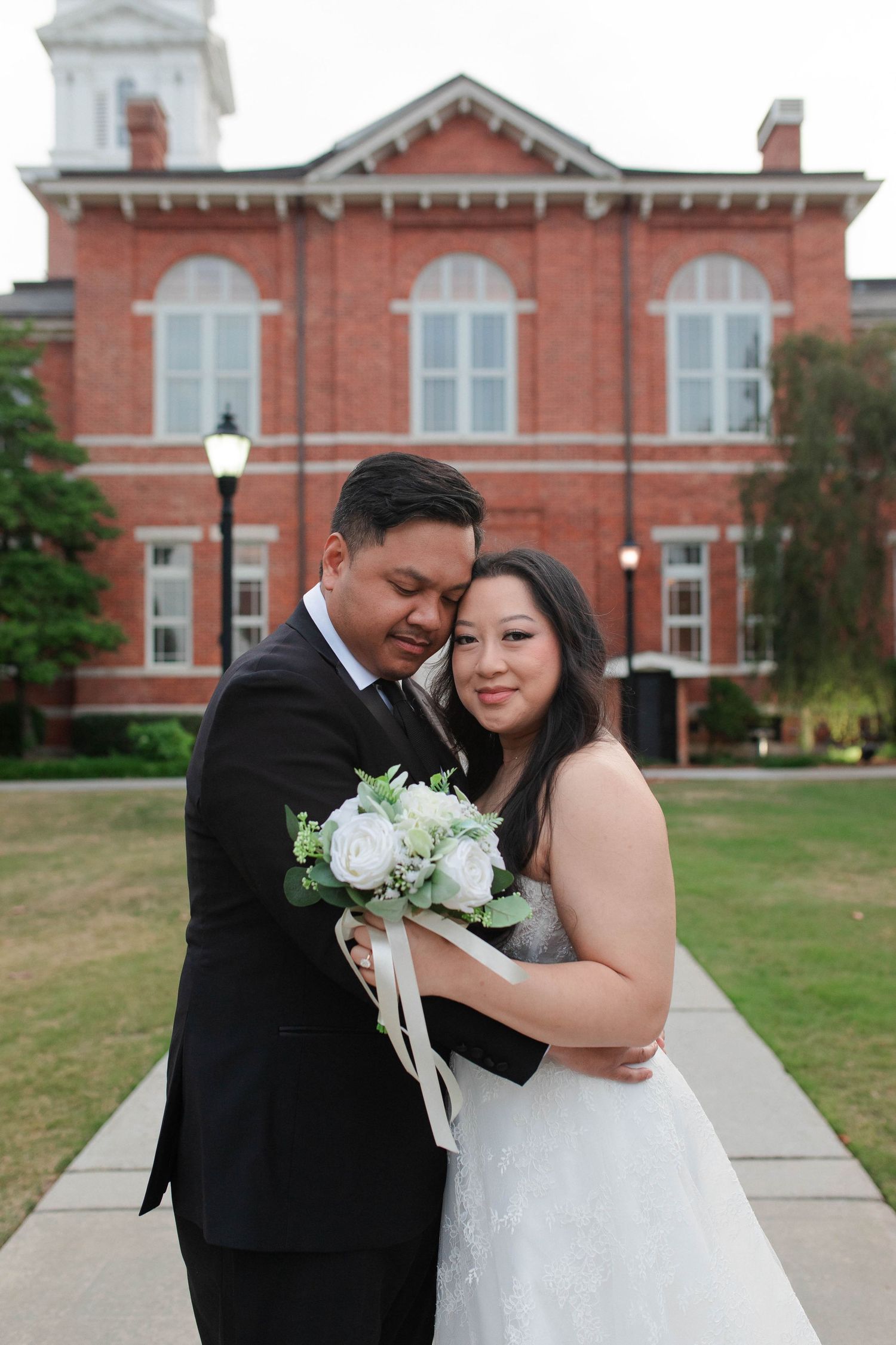 Bride and groom embrace outdoors in front of a red brick building, holding a white floral bouquet.