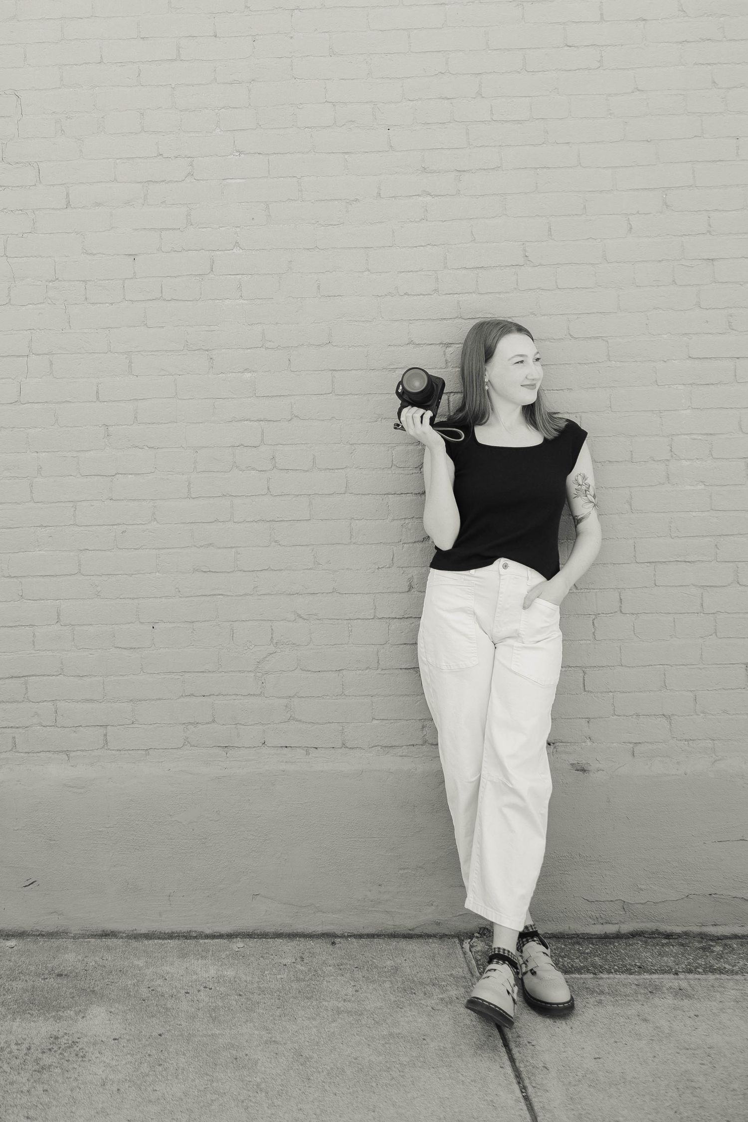 Black and white photo of a young woman holding a camera, leaning against a brick wall in white pants and black top.