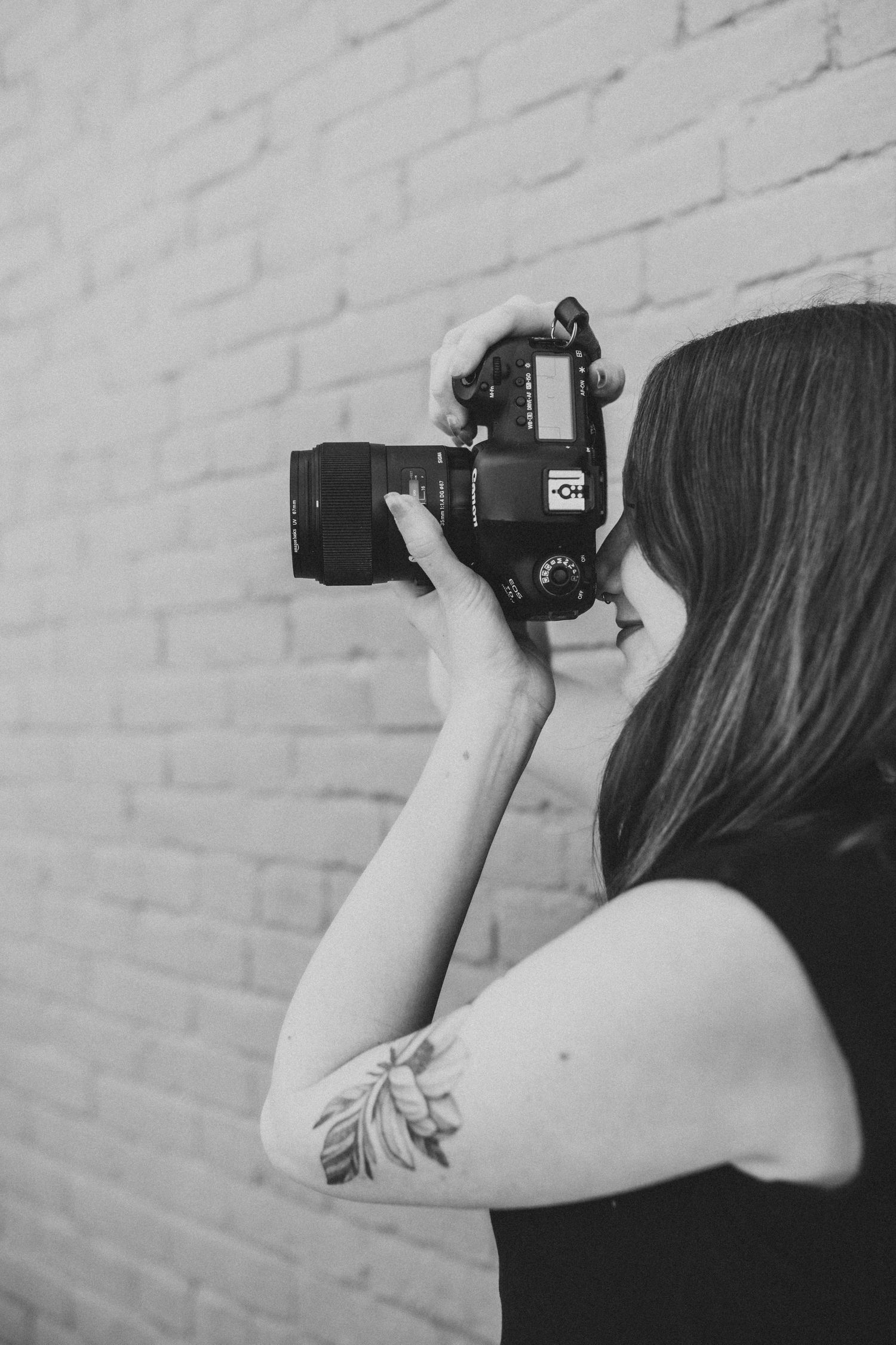 Black and white photo of a woman with a floral arm tattoo holding a DSLR camera up to her face near a brick wall.