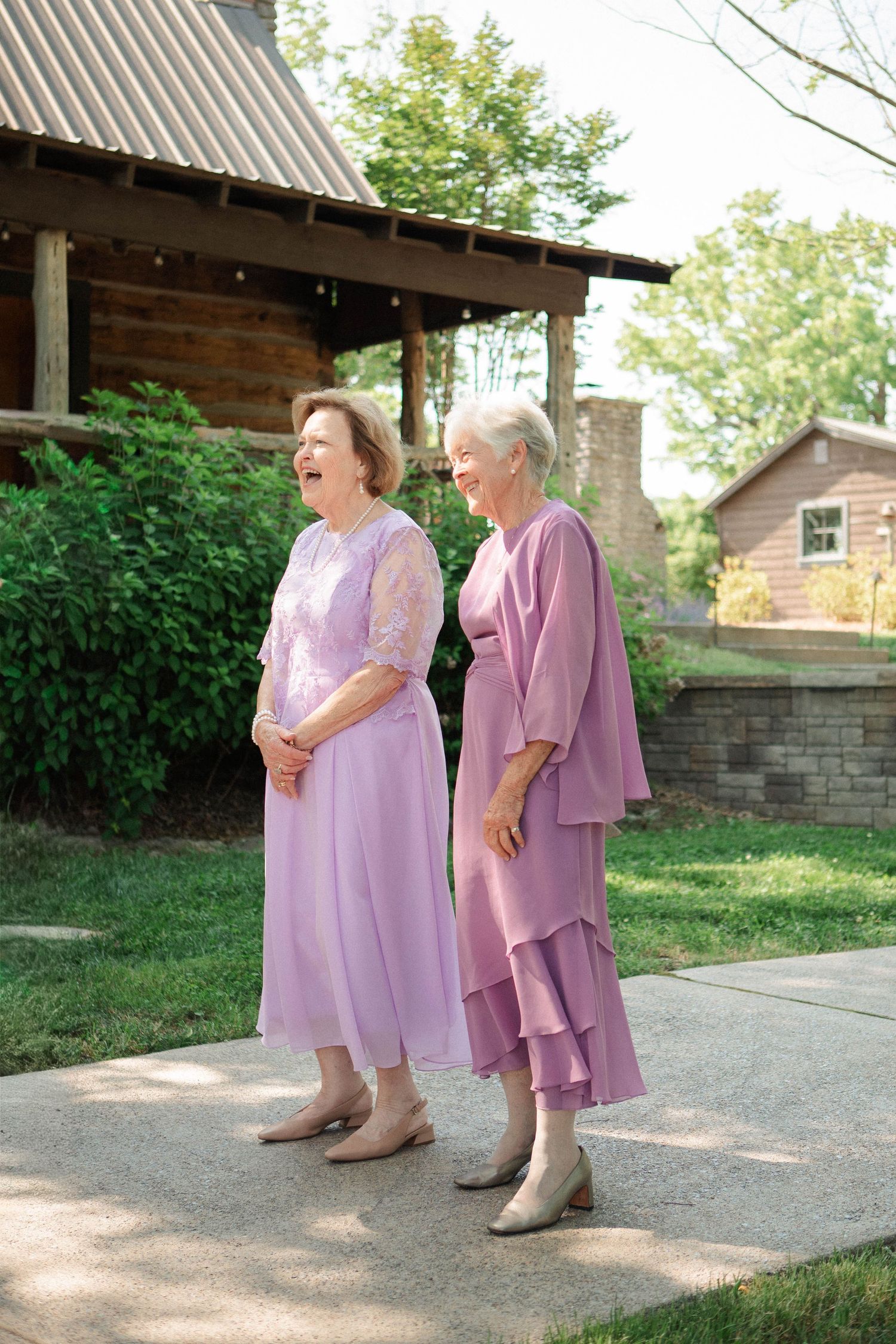 Two elegant senior women in matching lavender dresses laughing together outside a rustic log building.