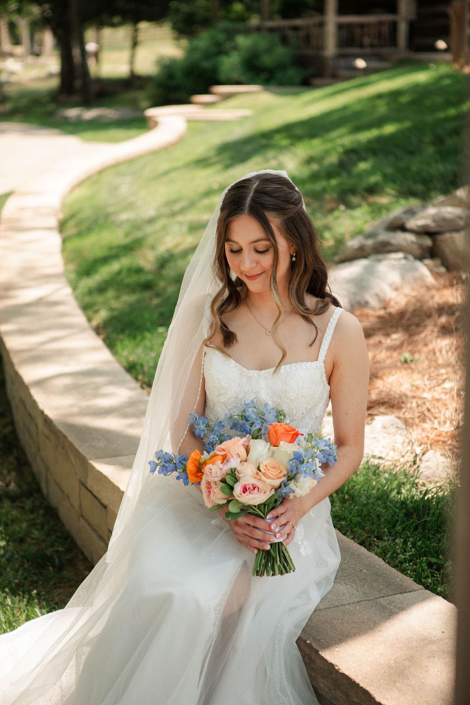 Bride in white gown holds colorful bouquet of blue and orange flowers on stone steps outdoors.