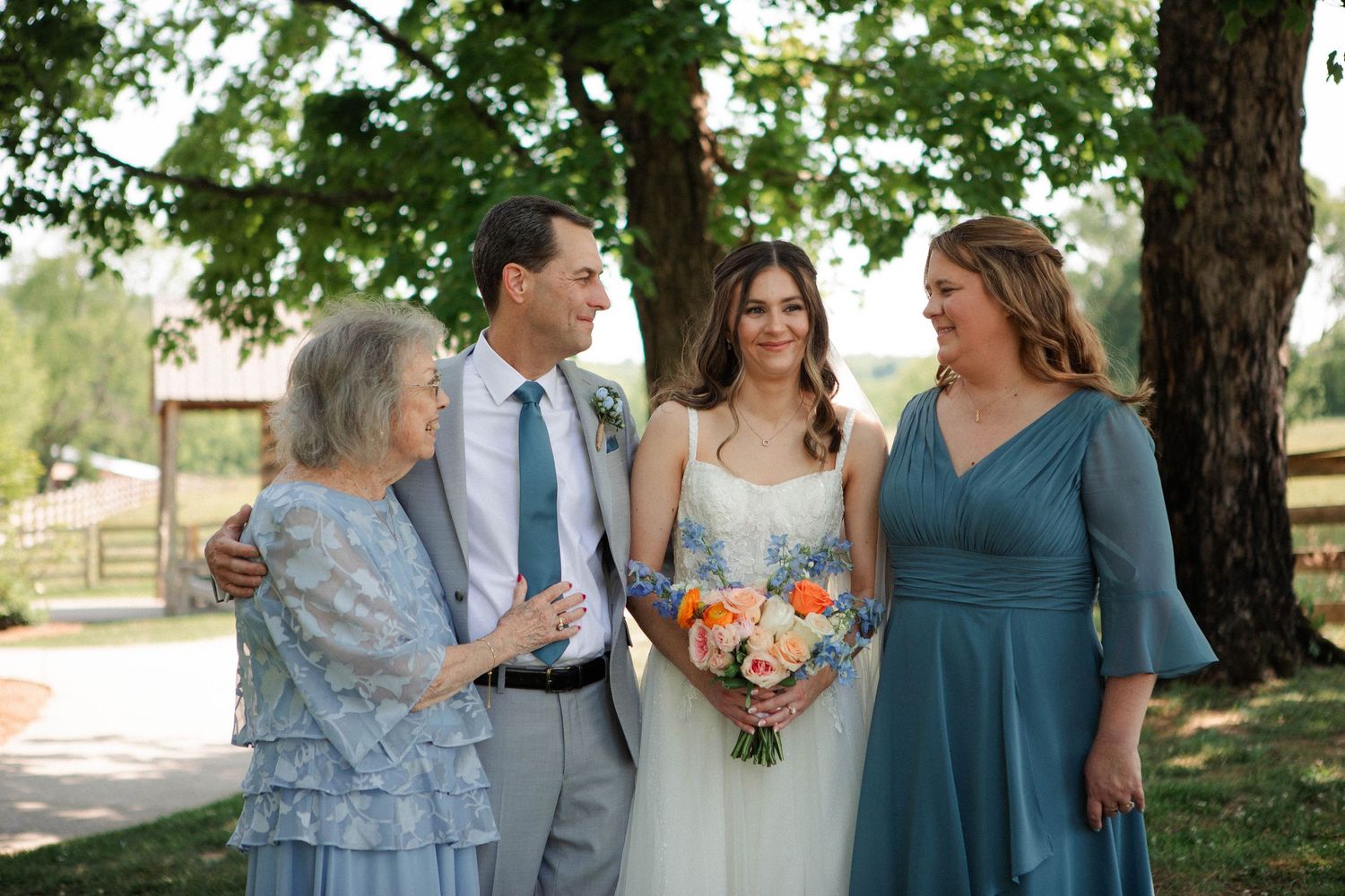 Bride holding colorful bouquet poses with family members in teal and blue dresses outdoors under trees.