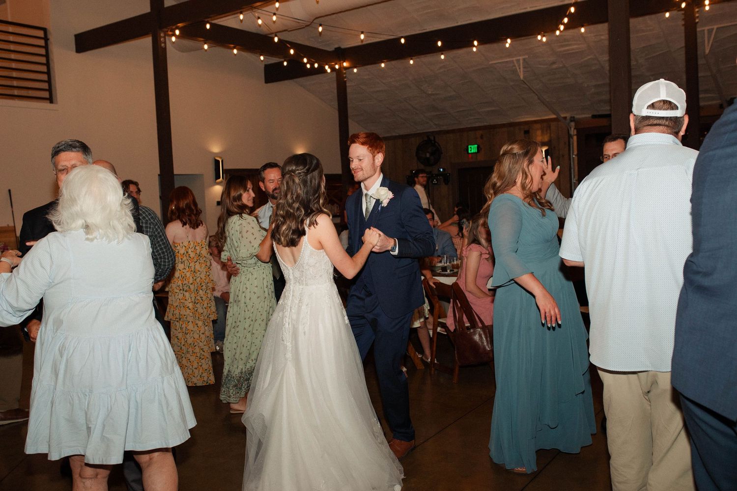 Bride and groom share a dance surrounded by guests at a rustic wedding reception venue with string lights.