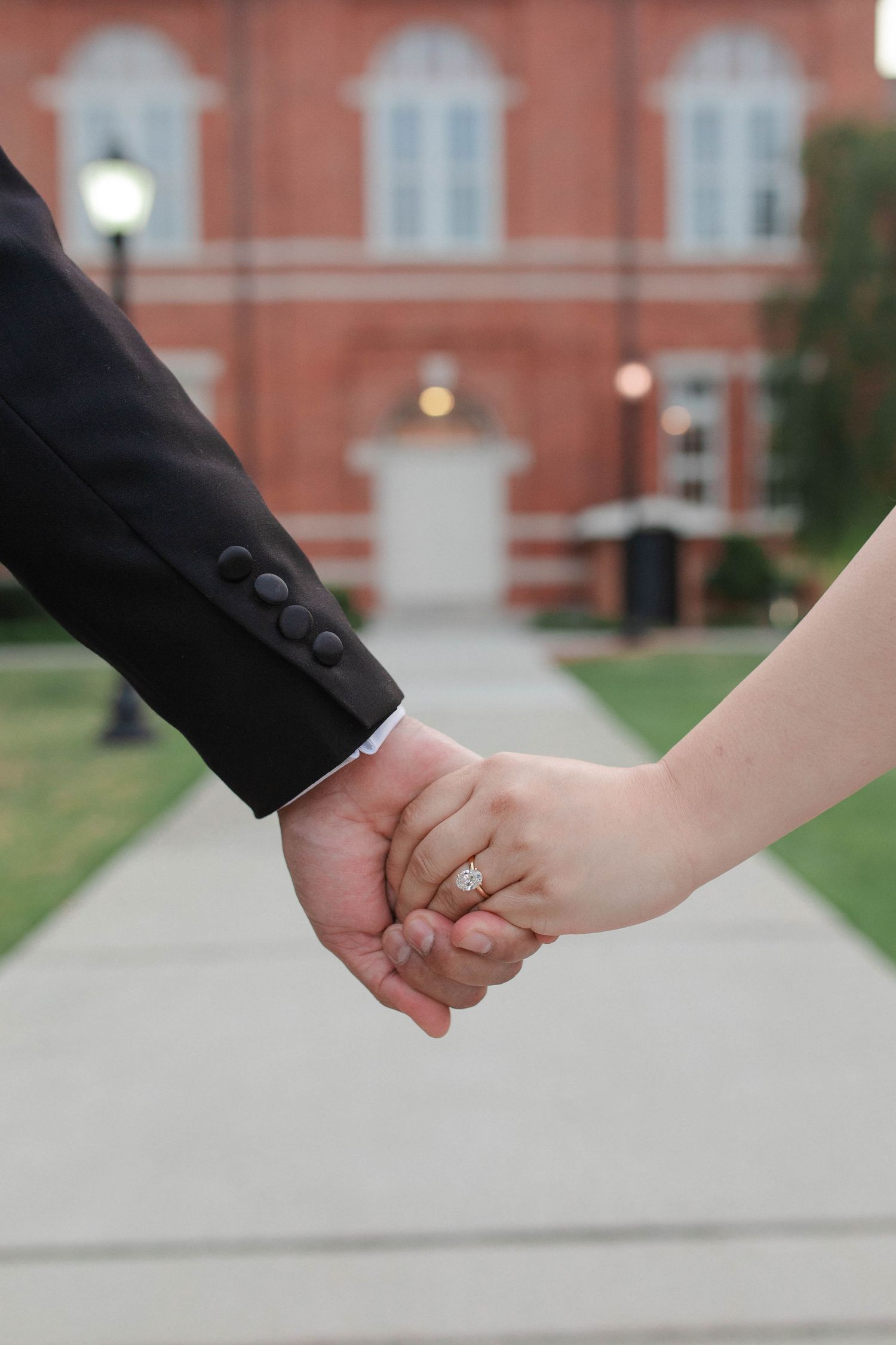 Couple holding hands outdoors, woman wearing gold engagement ring, man in black suit jacket, red brick building background.