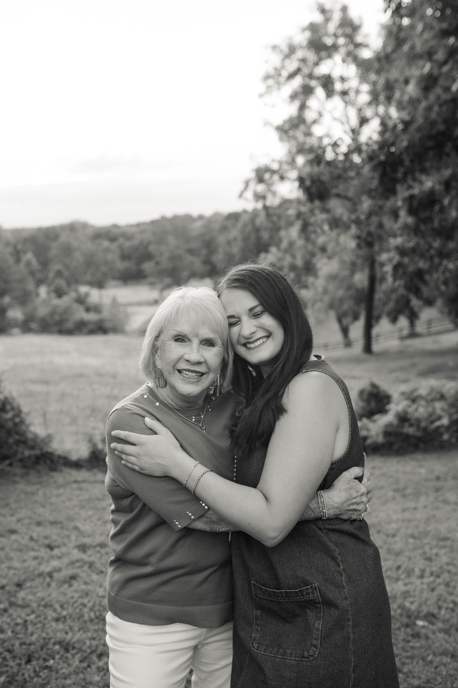 Black and white photo of two women hugging and smiling outdoors in a lush green park setting.