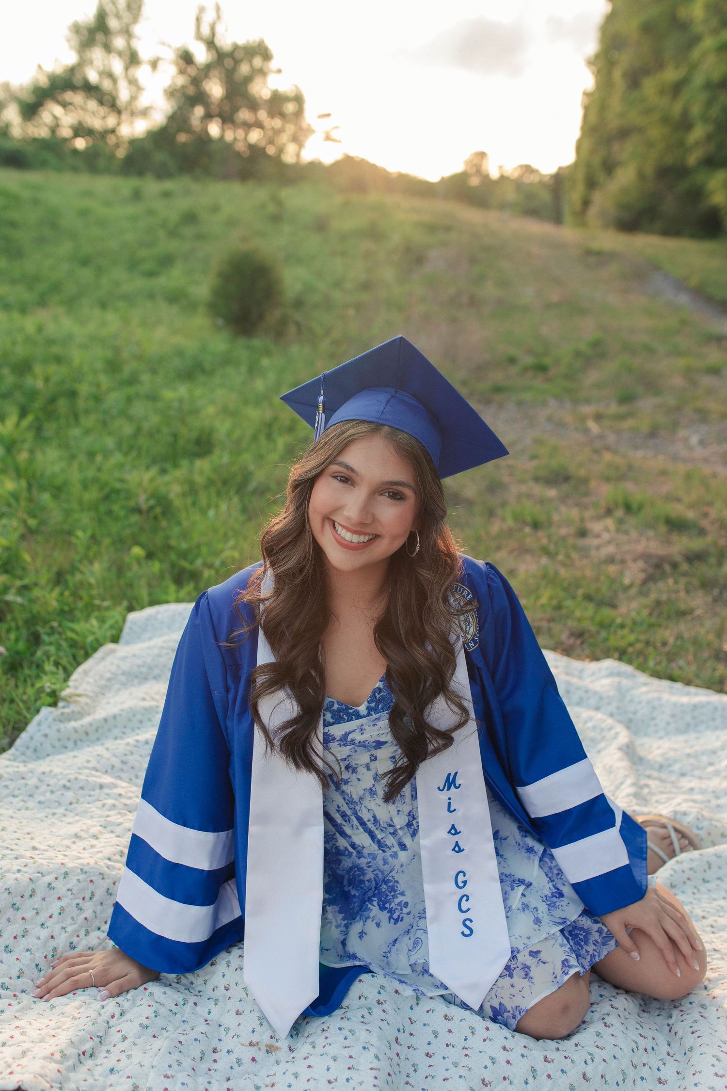 Smiling young woman in blue graduation cap and gown sits on blanket in green field at sunset wearing a white sash.