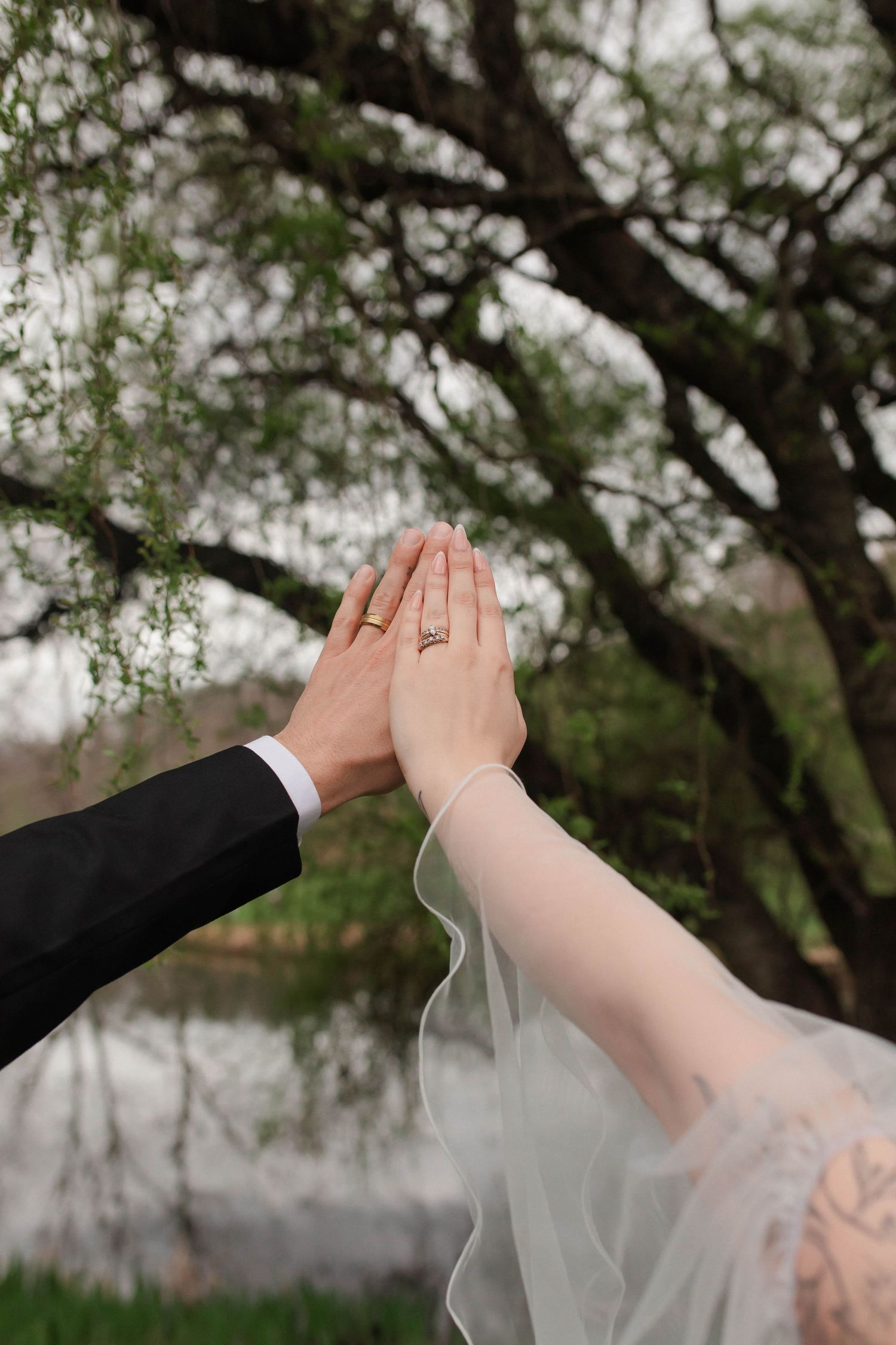Newlyweds press palms together showing wedding rings near a serene pond with lush green trees after eloping in Nashville.
