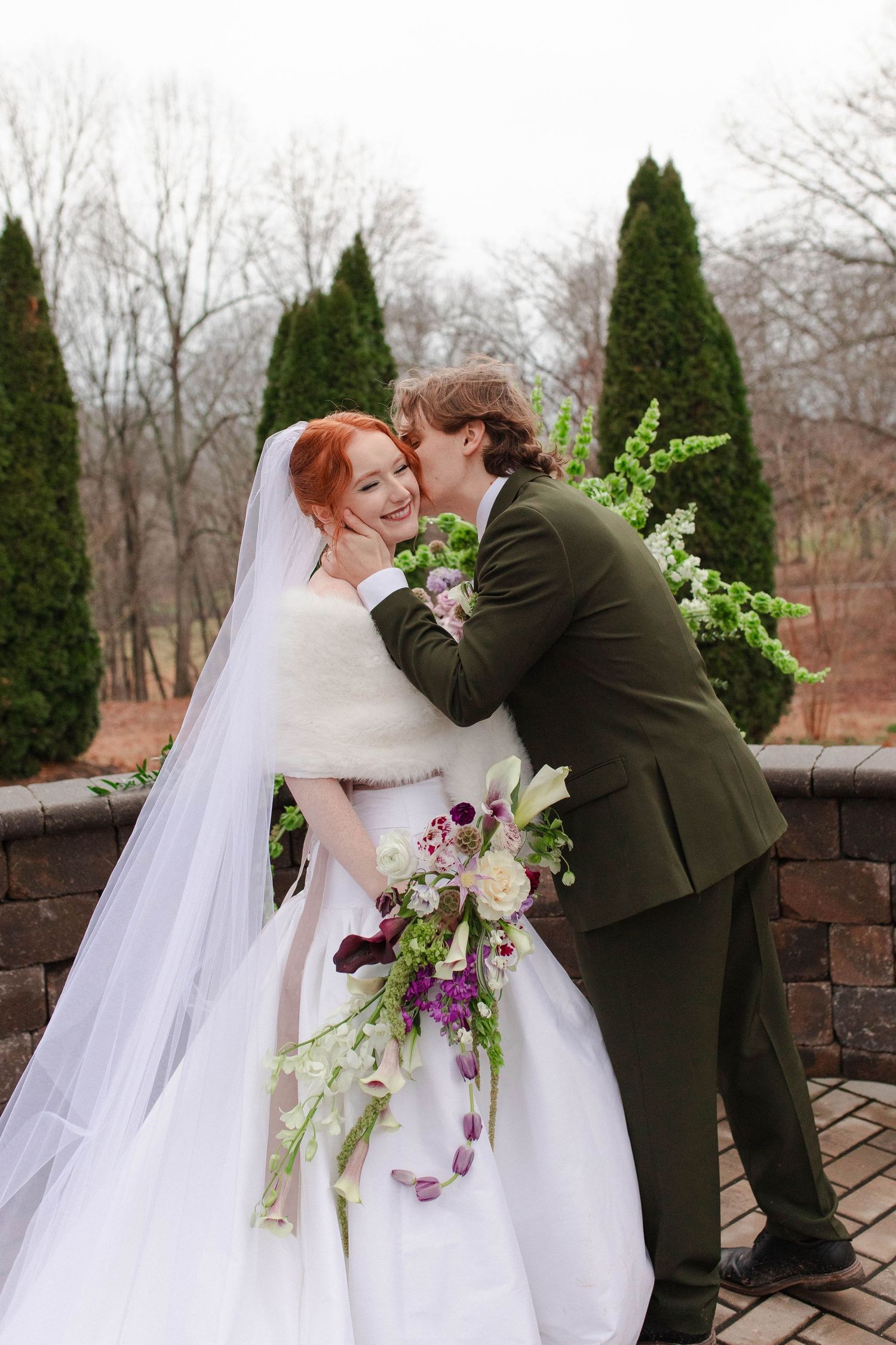 Groom in olive suit kisses red-haired bride on cheek while she holds cascading purple floral bouquet after eloping in Nashville.