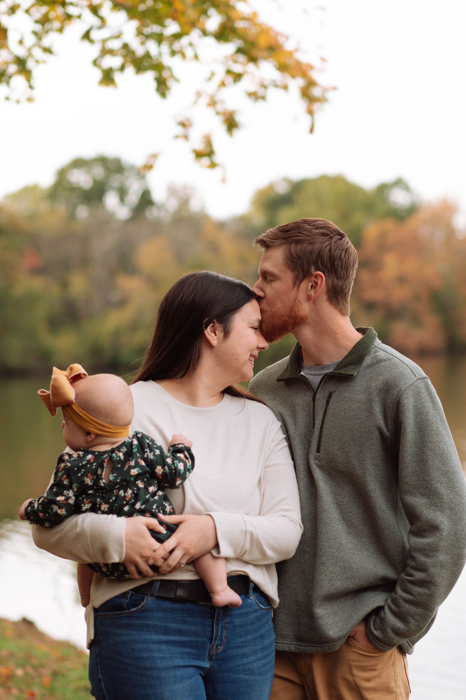 A couple shares a tender moment by a lake in autumn, as the man kisses the woman's forehead while she holds their baby.