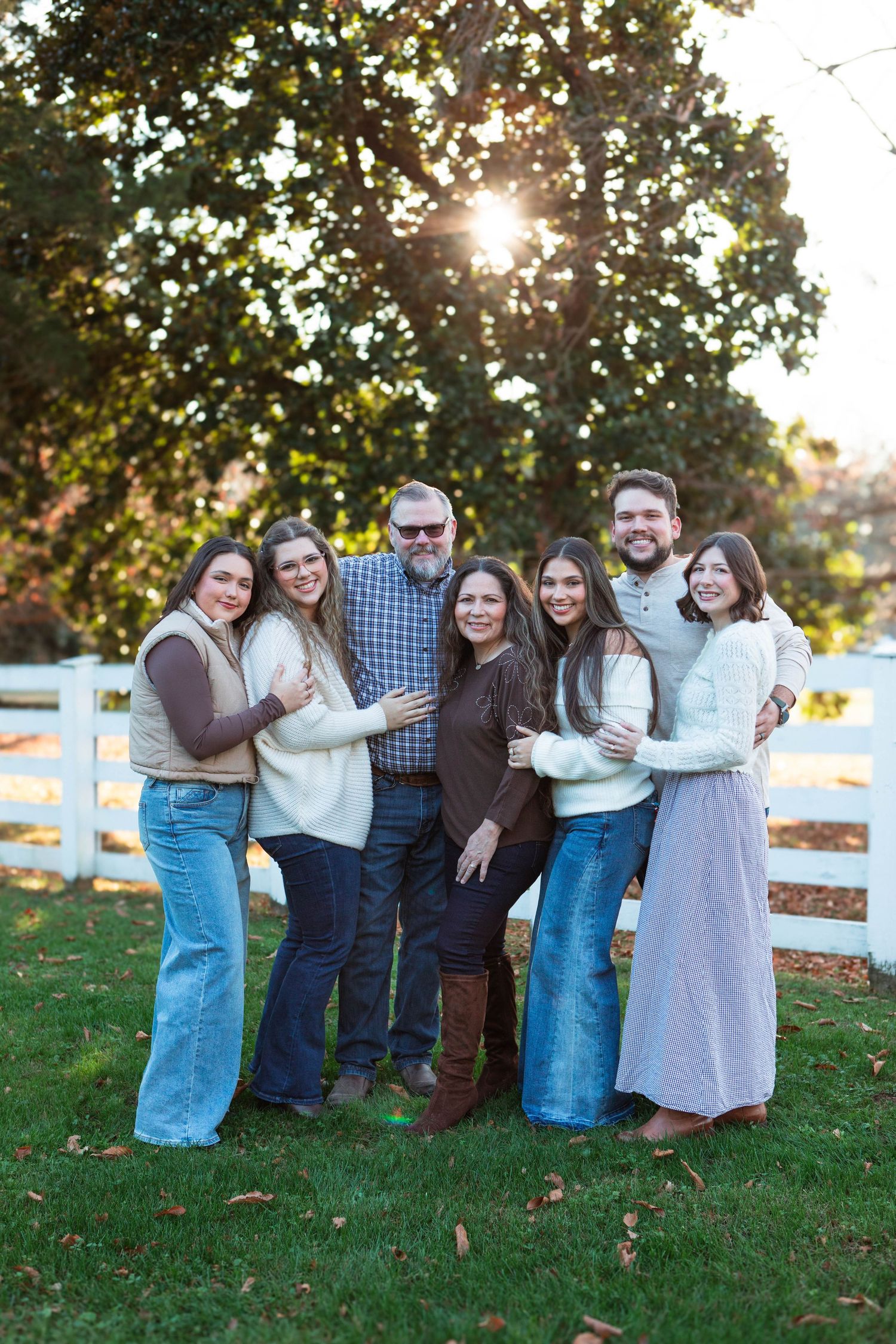 A joyful family of seven poses outdoors near a white fence and large tree with sunlight filtering through.